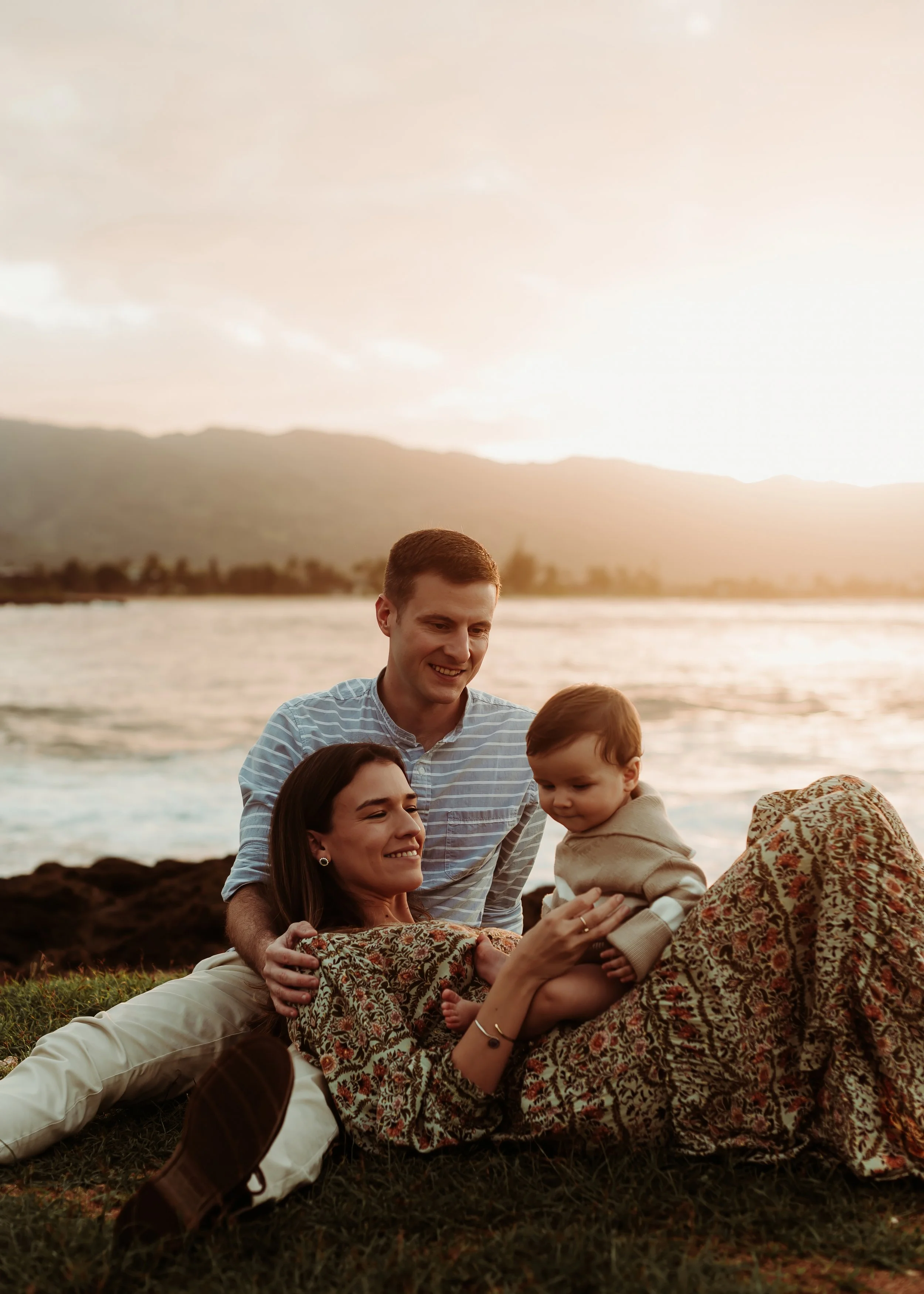 A family enjoying a sunset on the beach, sitting and playing on the grass with ocean and mountains in the background