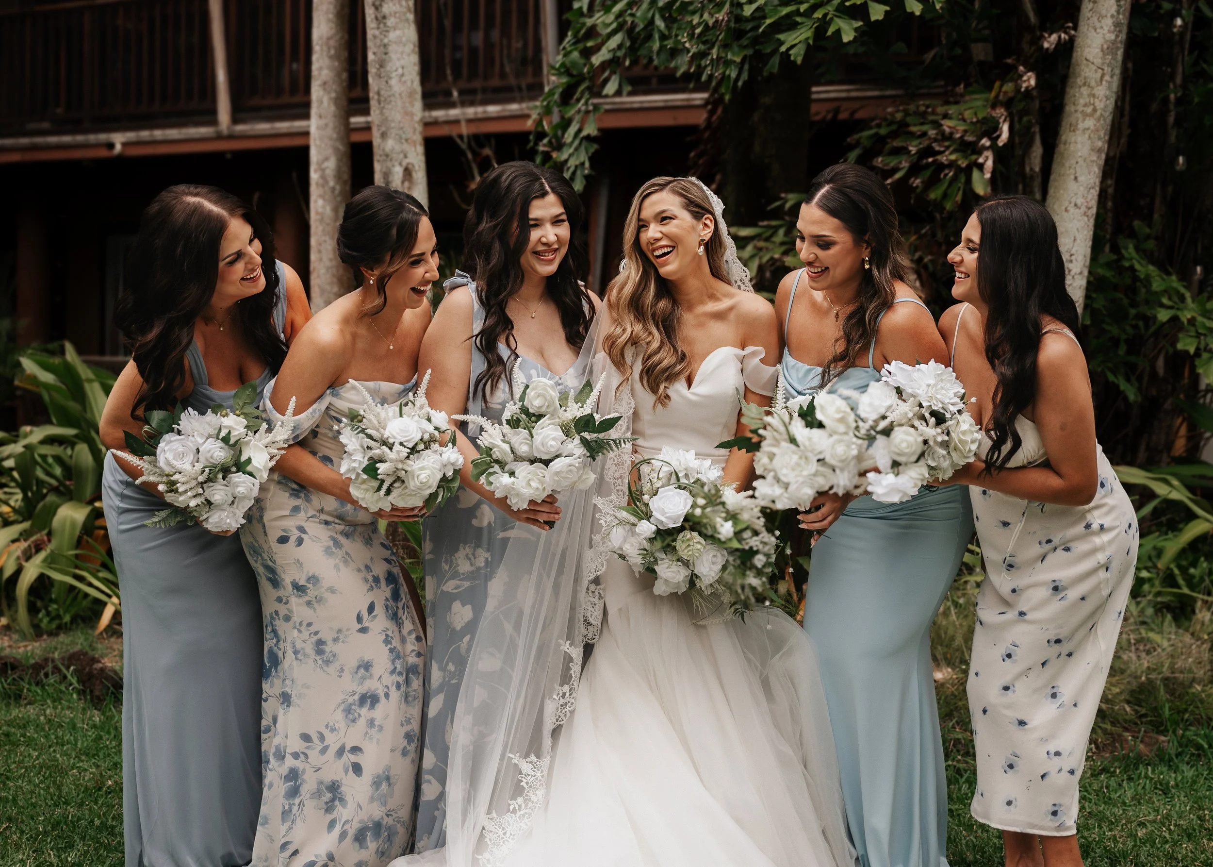 A bride in a wedding dress and veil surrounded by six bridesmaids in blue floral dresses holding white bouquets, standing outdoors with trees in the background, all smiling and laughing. | Best Hawaii Wedding Photographer & Videographer | Oahu, Hawai