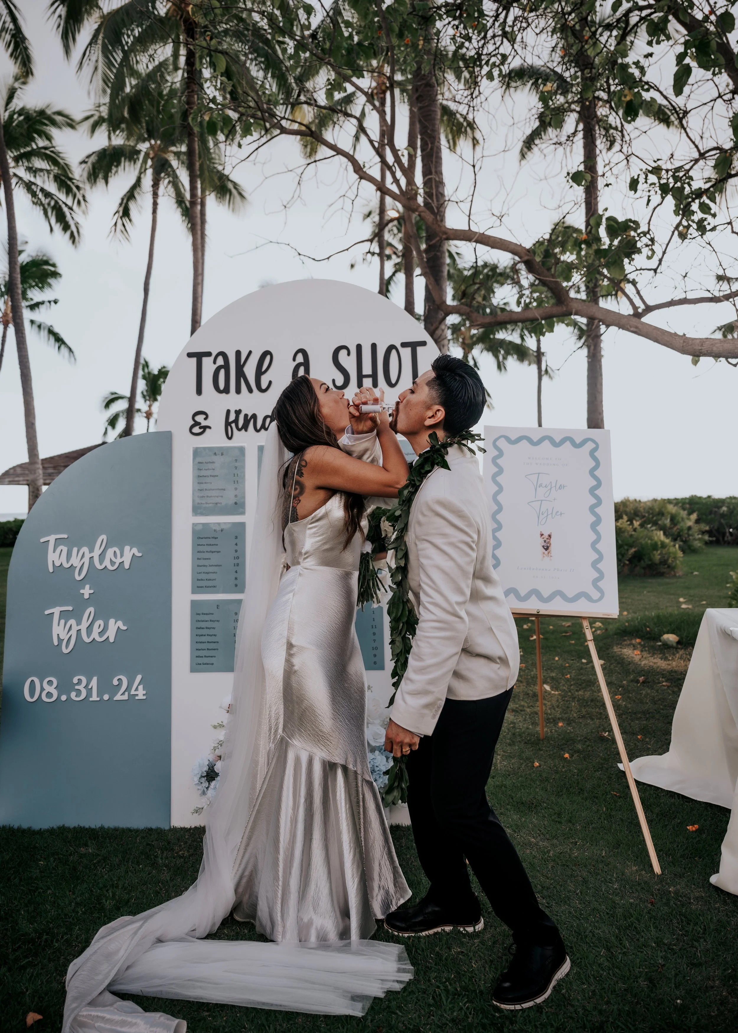 A bride and groom share a kiss at their wedding, standing outdoors in front of a sign that reads "Take a Shot & find" with a menu, and greenery and palm trees in the background. The bride is holding a shot glass up to the groom's mouth, and they are 