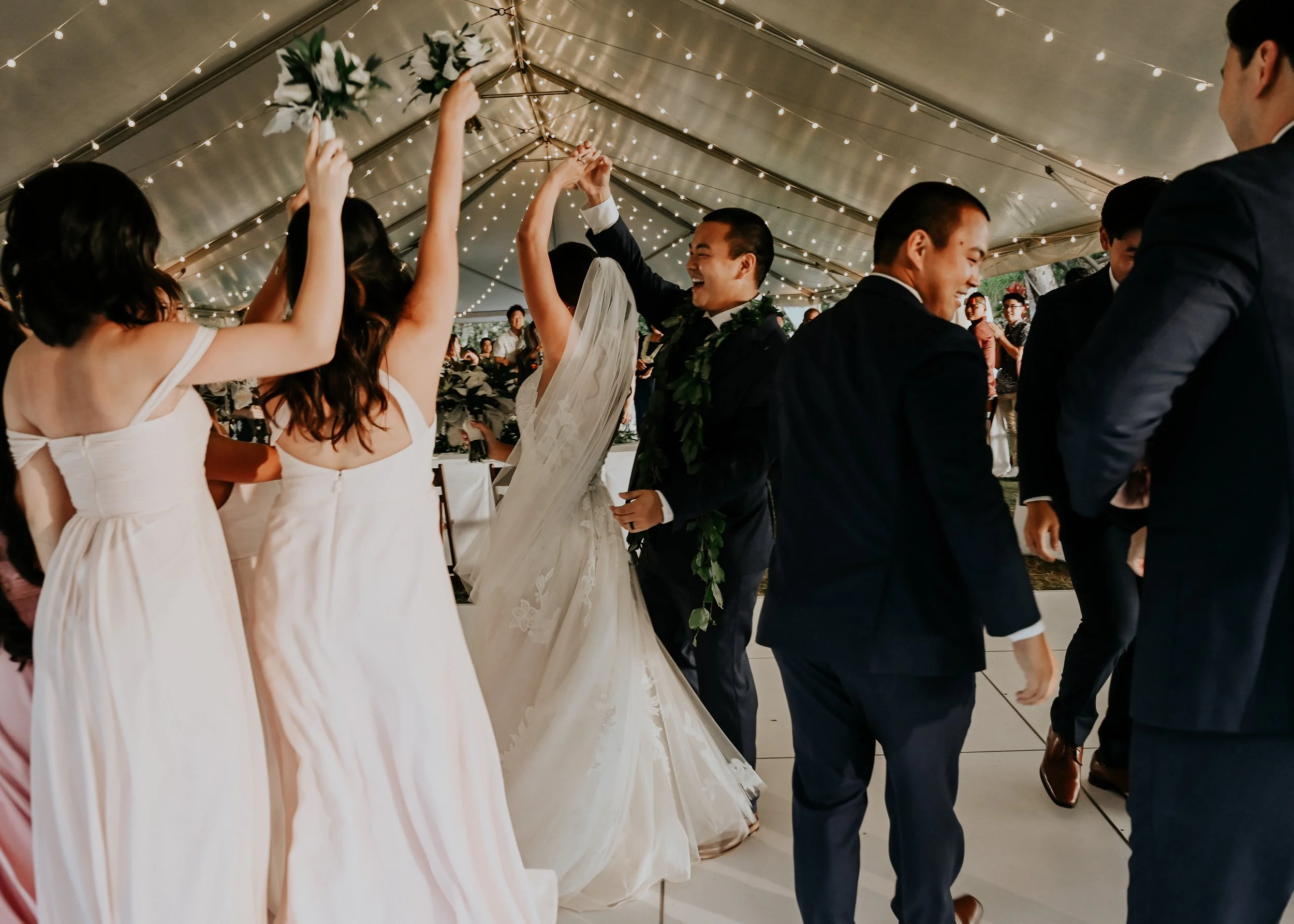 A bride and groom and their wedding party at a wedding reception under a tent decorated with string lights, with a bride and groom holding hands in the center. | Best Hawaii Wedding Photographer & Videographer | Oahu, Hawaii Island, Kauai, and Maui 