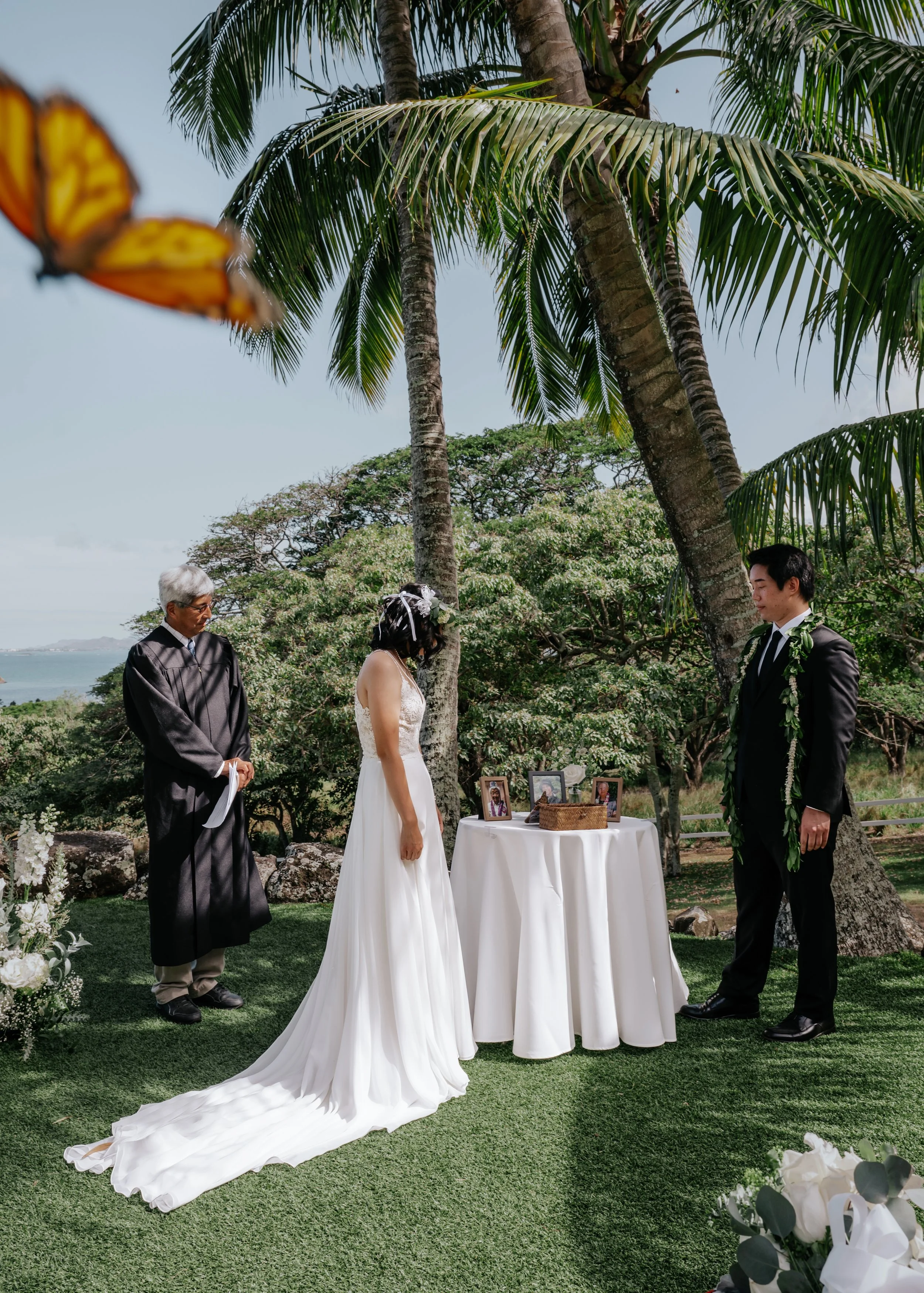 A wedding ceremony is taking place outdoors under tall palm treesThe bride, wearing a white gown with a long train, is standing opposite the groom, who is dressed in a black suit with a green lei. An officiant, dressed in a black robe and glasses, stands to the side. A small table covered with a white cloth, holding framed photos and decorations, is placed between the couple. Floral arrangements are visible in the foreground.