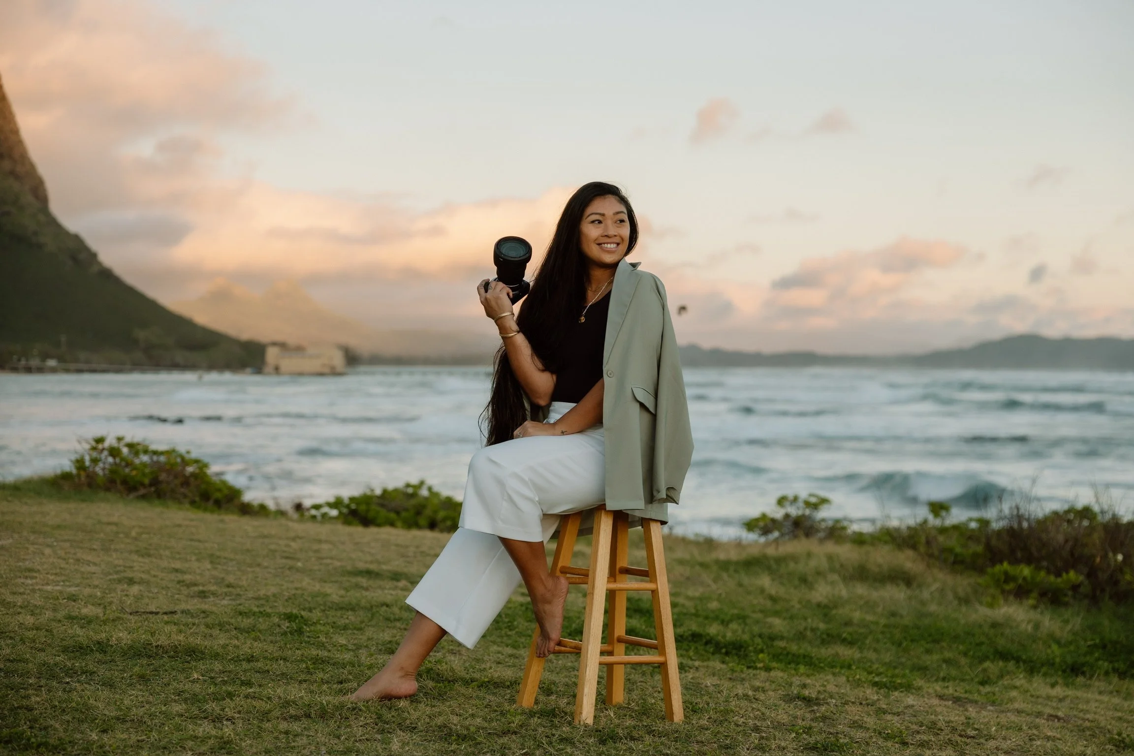 A woman, the owner Ashley Gallarde with long dark hair sitting on a stool near the beach at sunset, holding a camera, smiling, wearing a black top, light green blazer, and white pants, with a scenic ocean and mountain background.