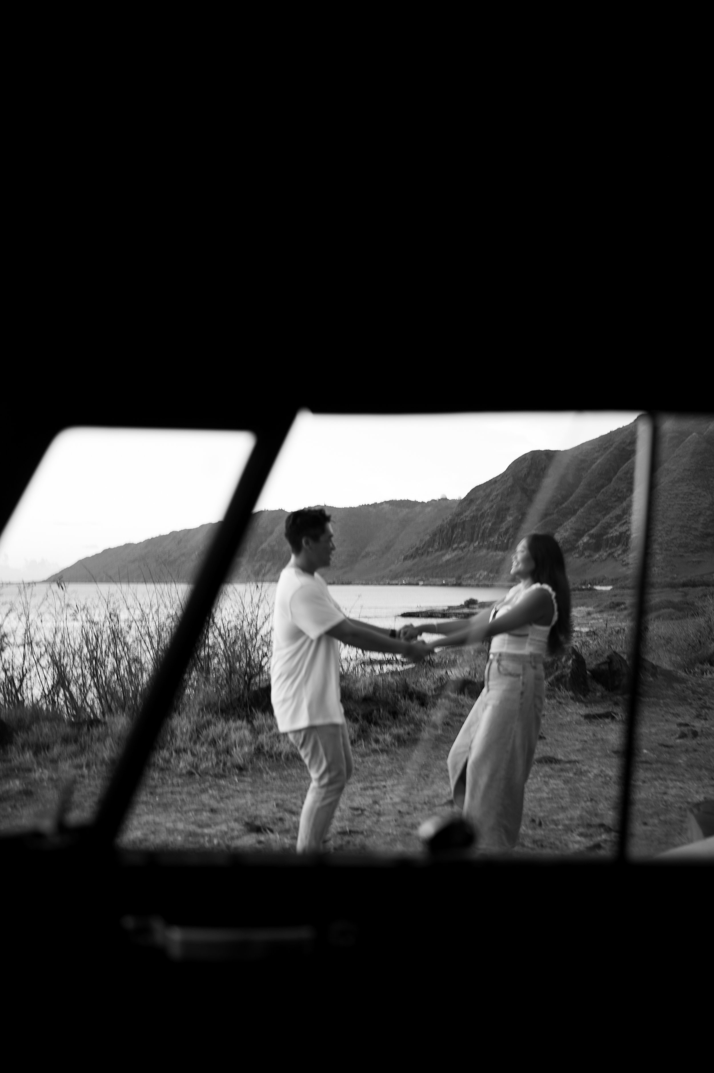 A black and white photograph of a couple standing by the beach in Hawaii, holding hands and facing each other, with mountains in the background.  | Best Hawaii Wedding Photographer & Videographer | Oahu, Hawaii Island, Kauai, and Maui