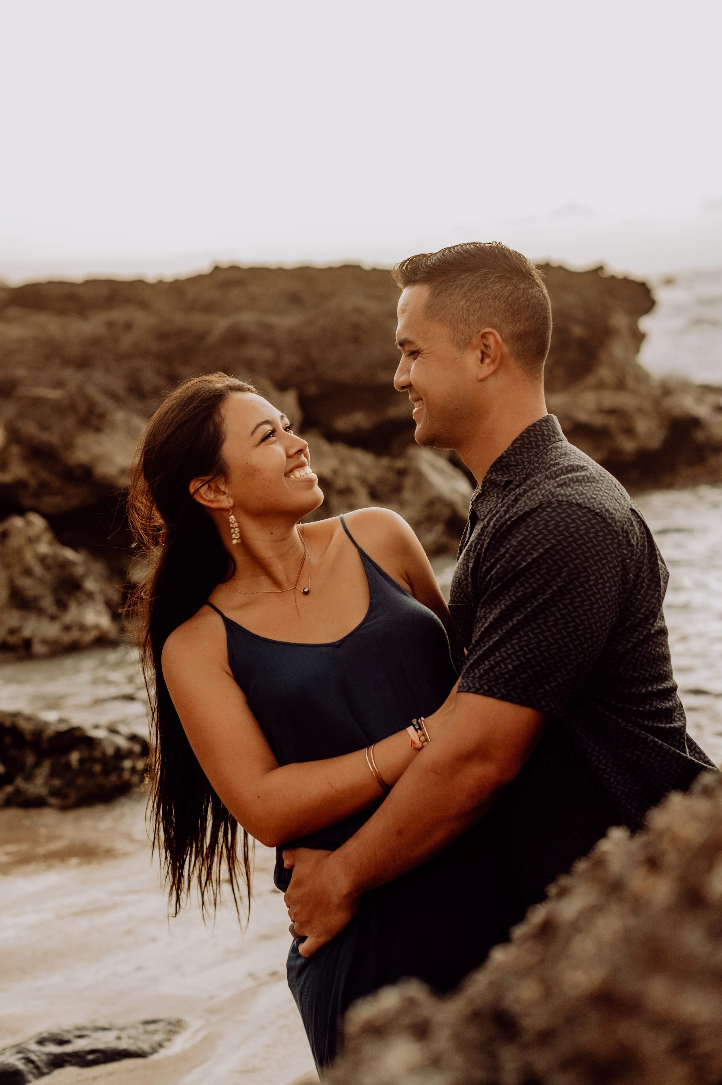 A couple smiling and looking at each other on a beach, with rocks and ocean in the background.