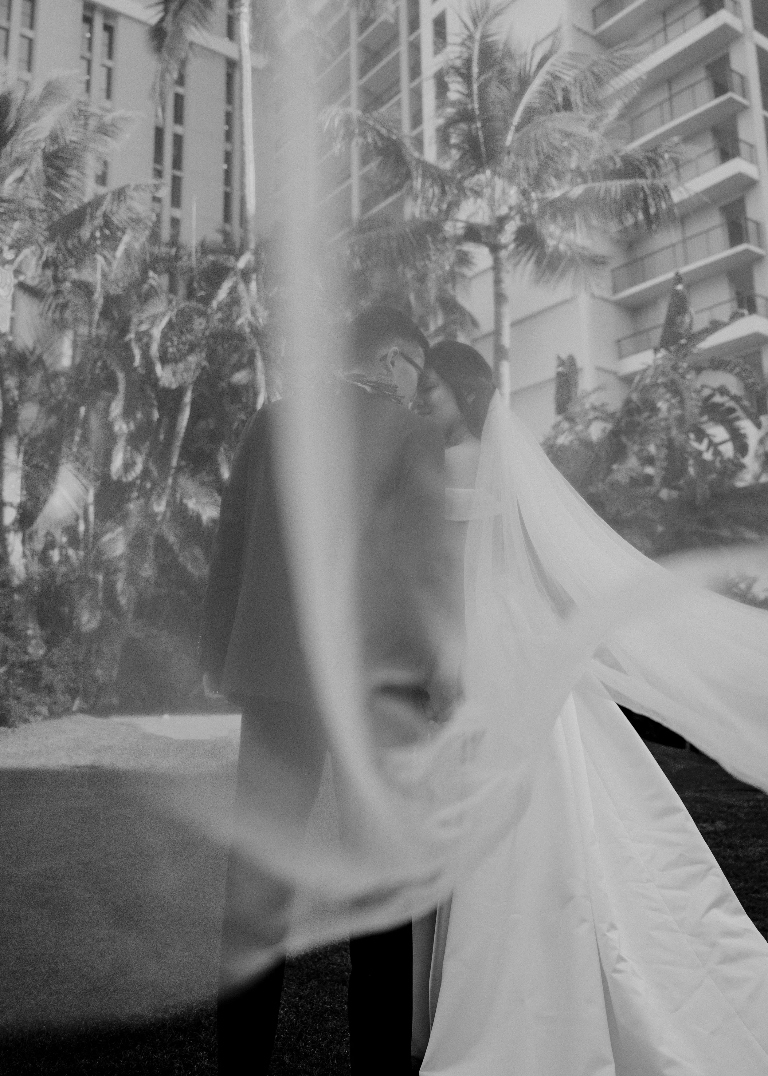 A side view of a bride and groom holding hands, standing outdoors near palm trees and a tall building. They are dressed in wedding attire, with the bride in a white gown and veil, and the groom in a dark suit.