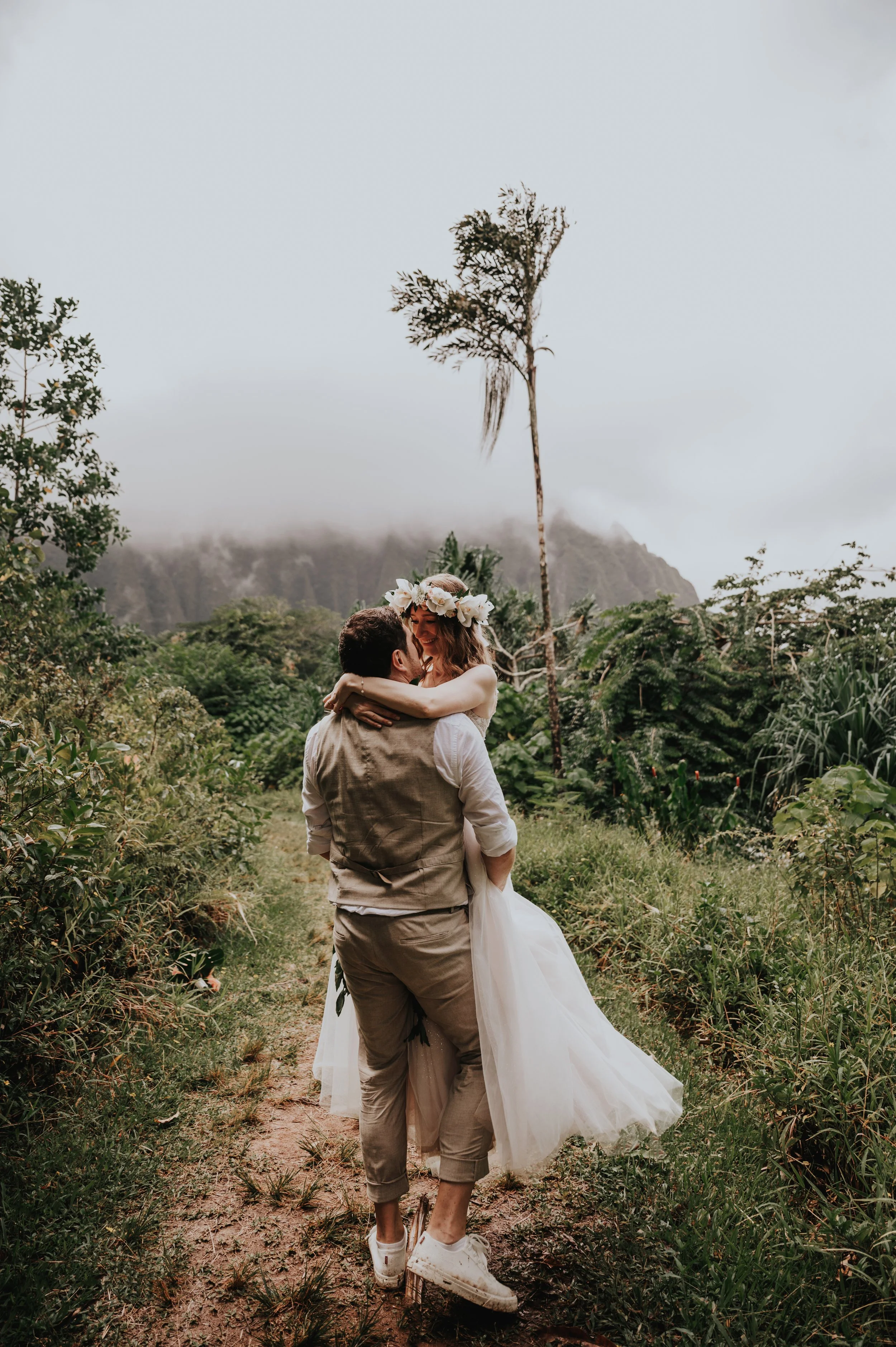 A couple dressed in wedding attire, with the woman in a white dress and floral crown, and the man in a beige vest, is hugging and smiling in a lush outdoor setting with greenery, a dirt path, mountain in the background, and overcast sky.