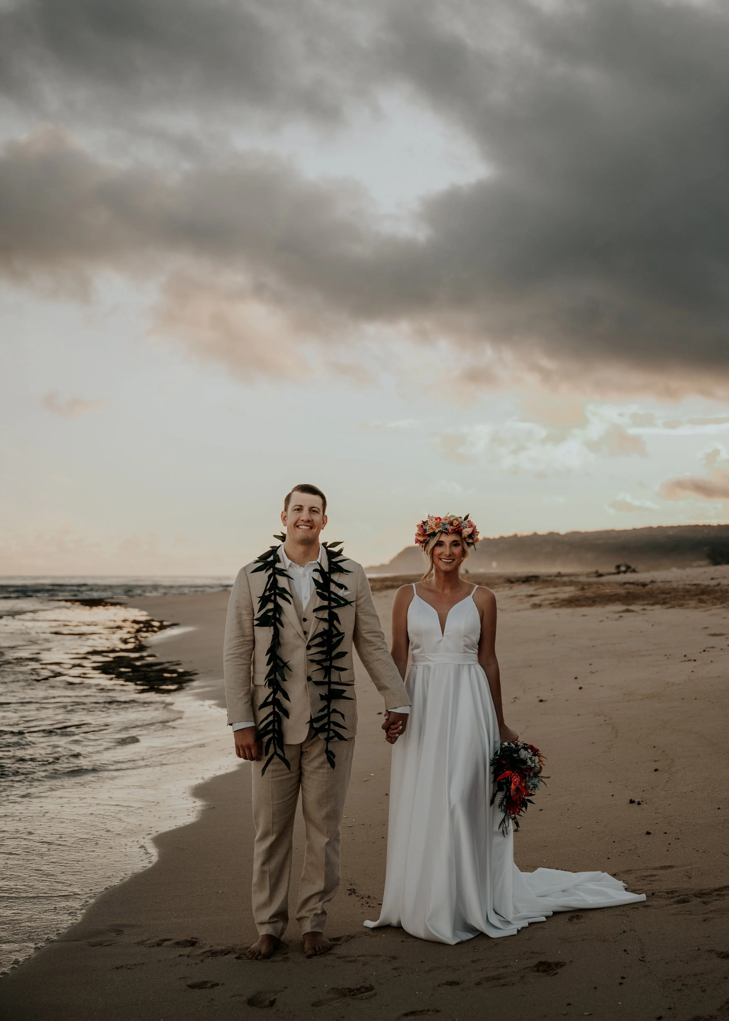 During and elopement a newly married couple holding hands and smiling on a beach at sunset. The groom is wearing a tan suit with a dark leafy lei, and the bride is wearing a white dress with a floral crown and holding a bouquet of flowers.