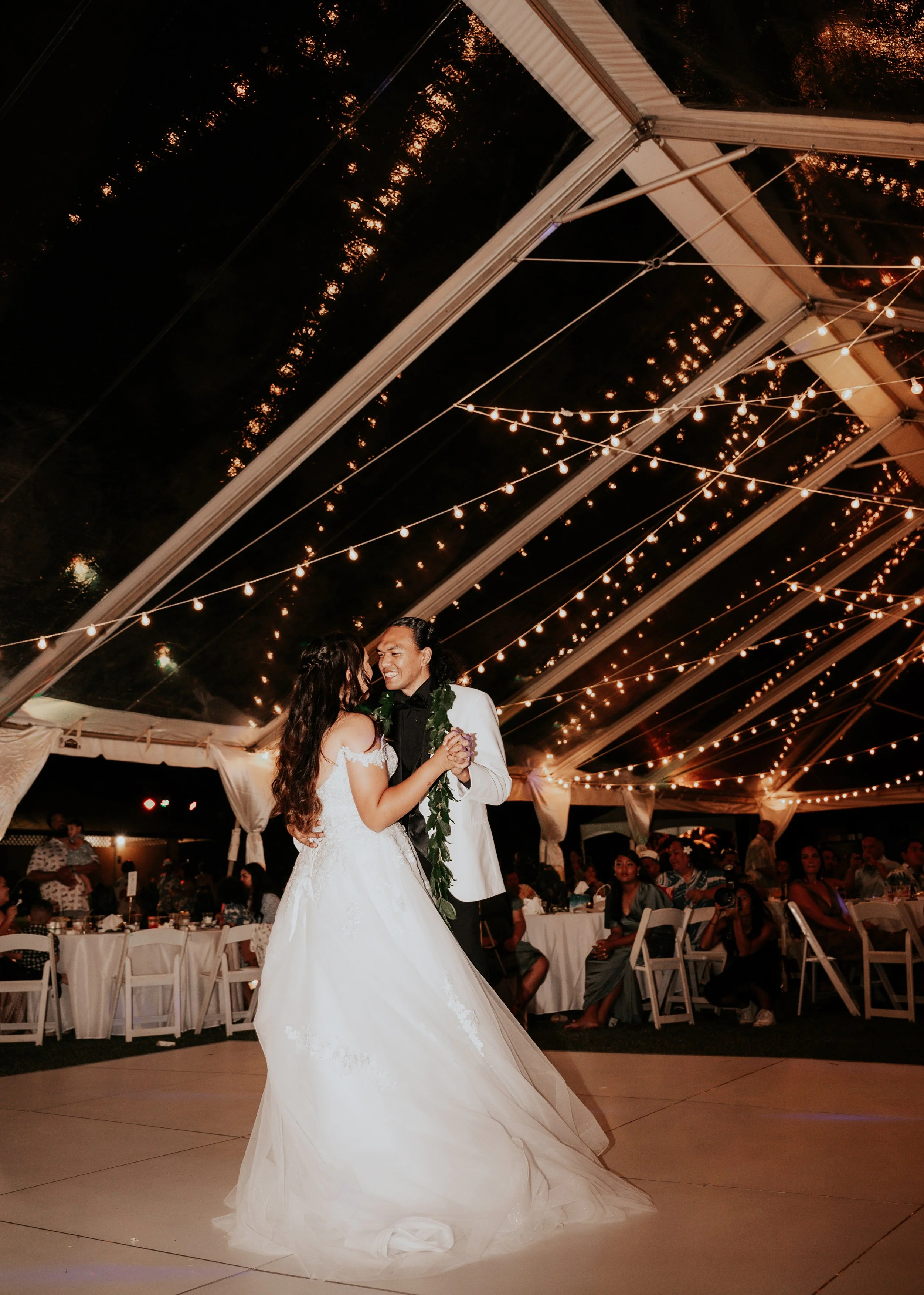 A bride and groom dancing at their wedding reception under a tent decorated with string lights at night, with seated guests watching in the background.