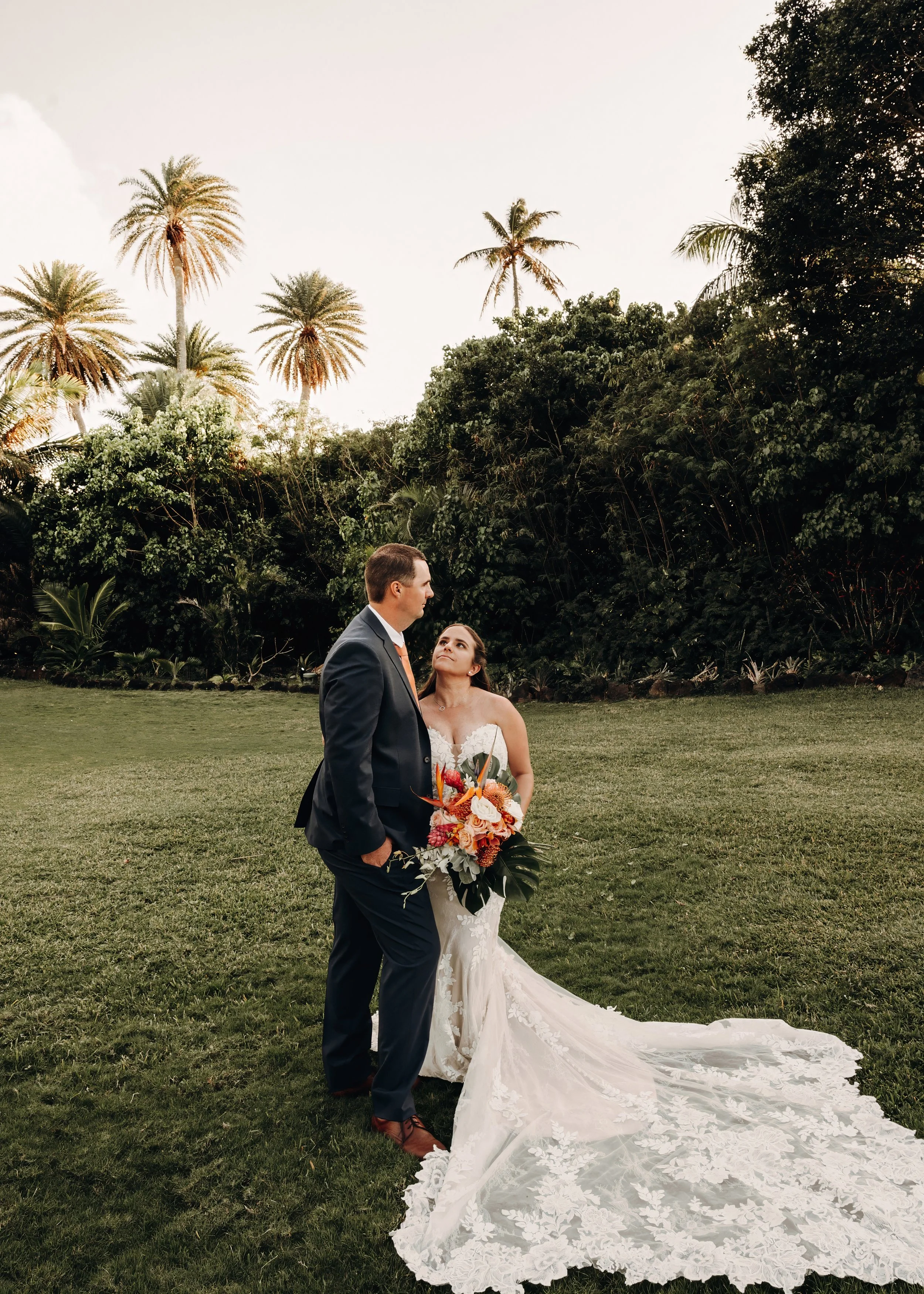 A bride and groom stand outdoors on grass, facing each other with trees and palm trees in the background. The bride holds a large bouquet of orange, red, and white flowers. She wears a white wedding gown with a long, lace train, and the groom is dressed in a dark suit and brown shoes. The sky is bright and slightly cloudy.