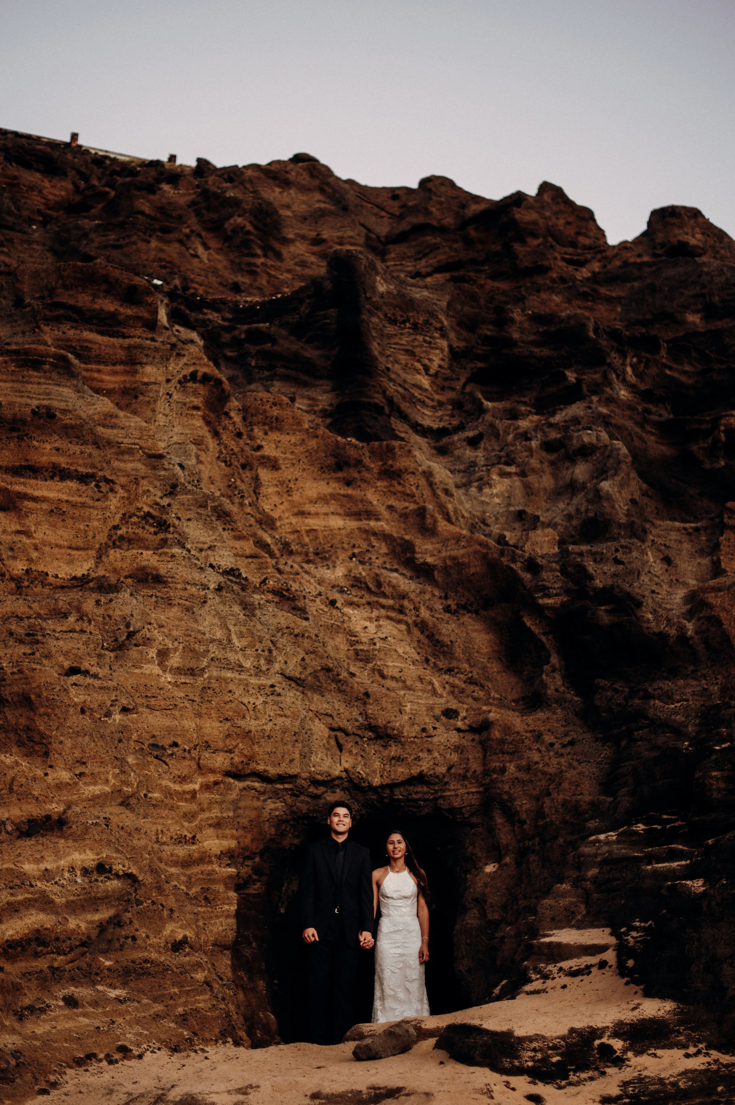 A man and woman in wedding attire standing together in front of a large rock formation, holding hands.
