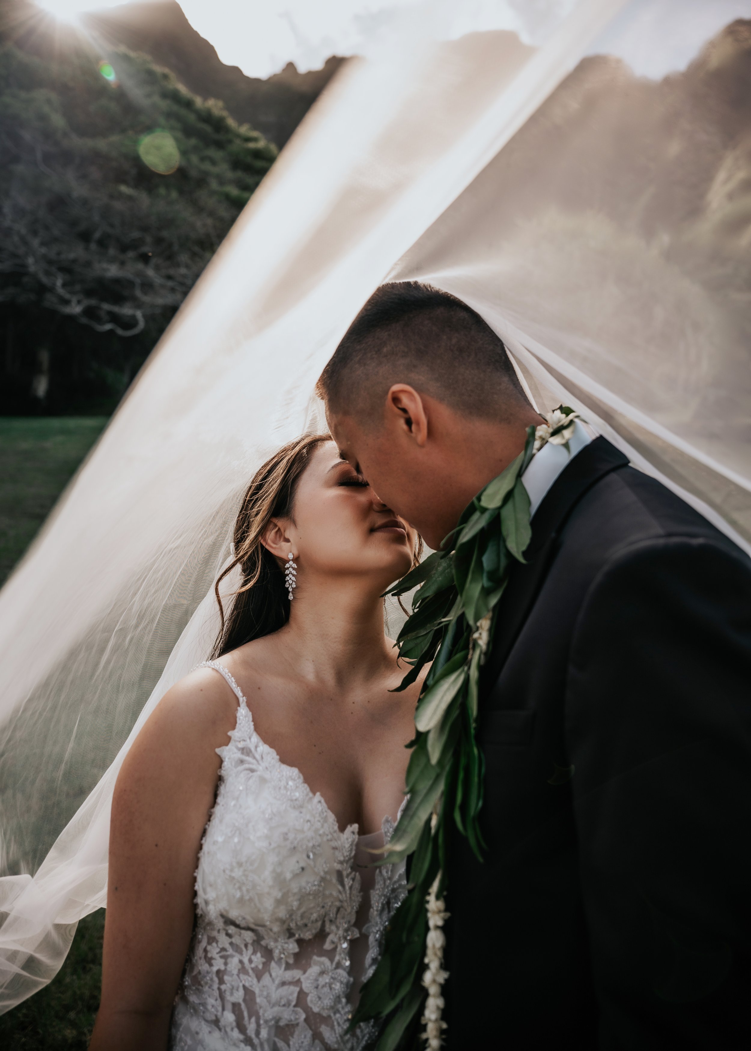A newlywed couple standing close with foreheads touching, under a wedding arch with mountains in the background.  | Best Hawaii Wedding Photographer & Videographer | Oahu, Hawaii Island, Kauai, and Maui