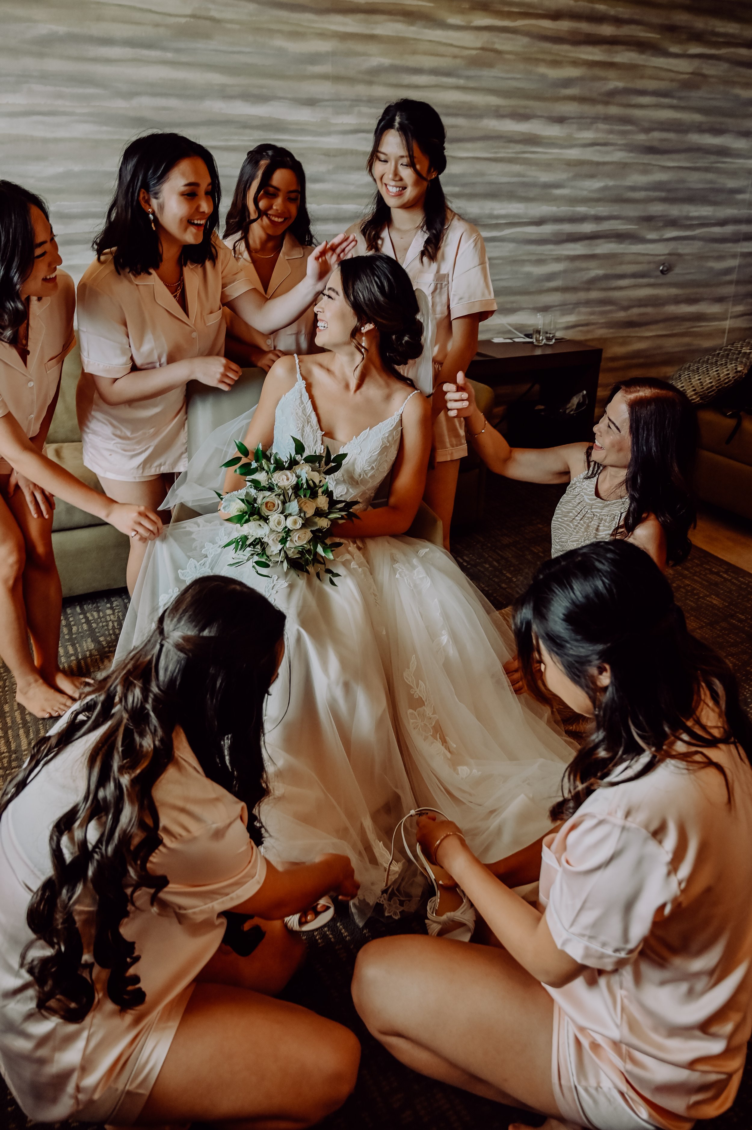 A bride with a bouquet of white roses surrounded by her bridesmaids in matching satin pajamas, some kneeling and others standing, inside a room with a wood-paneled wall.  | Best Hawaii Wedding Photographer & Videographer | Oahu, Hawaii Island, Kauai,
