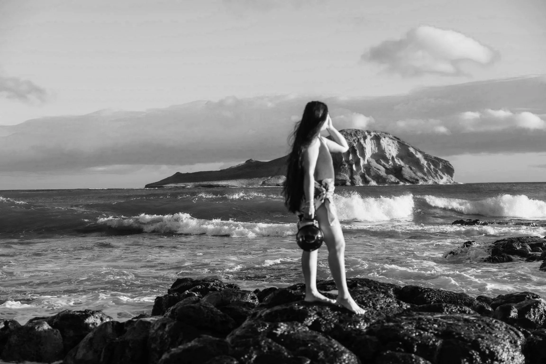 Woman with long hair standing on rocks by the ocean, with an island in the background, black and white photograph. | Wedding and Elopement Photography & Videography | Oahu, Kauai, Maui, Hawaii Island, Hawaii