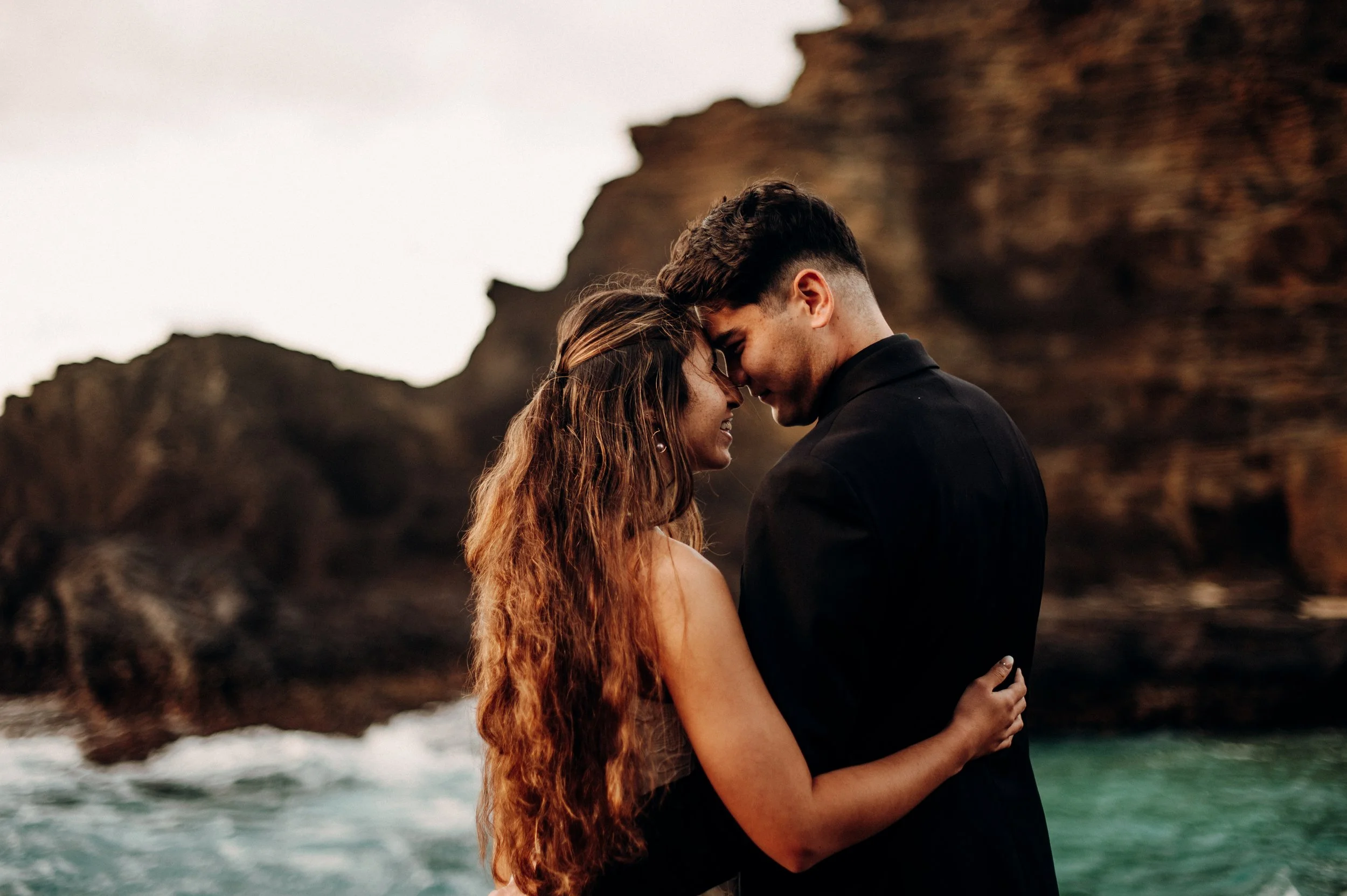 A couple stands close together with foreheads touching, smiling, on a rocky coastline with ocean waves and cliffs in the background at sunrise. | Best Hawaii Wedding Photographer & Videographer | Oahu, Hawaii Island, Kauai, and Maui 