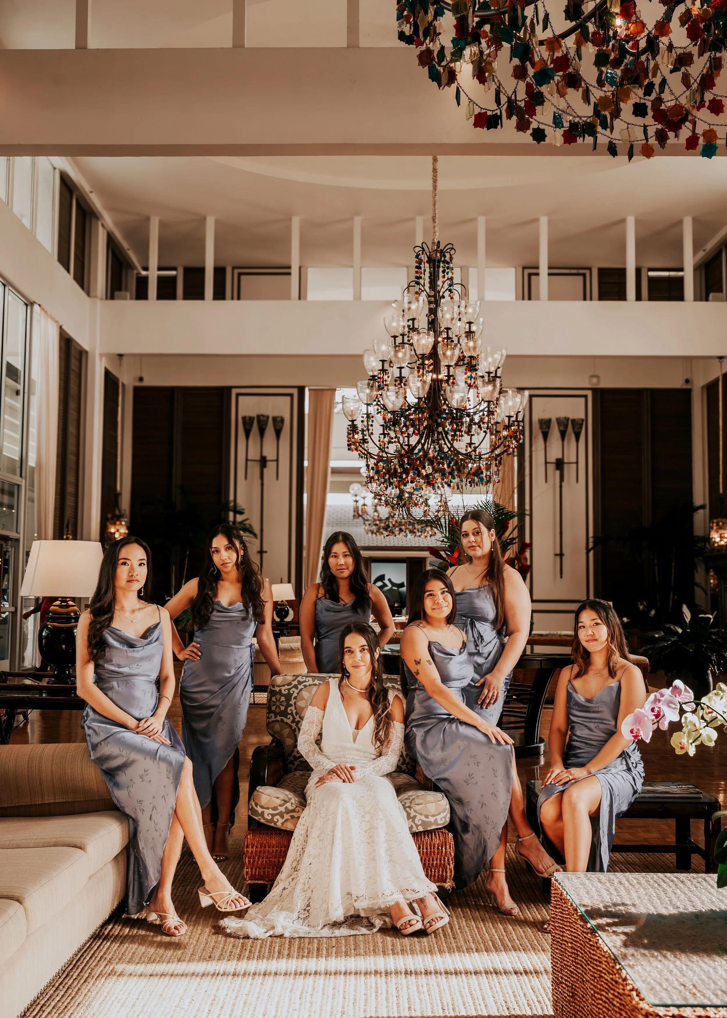 A bride and her bridesmaids dressed in matching satin dresses and high heels, pose in a luxurious hotel lobby decorated with a large chandelier and festive decorations. The bride, in a white lace wedding gown, is seated in the center.