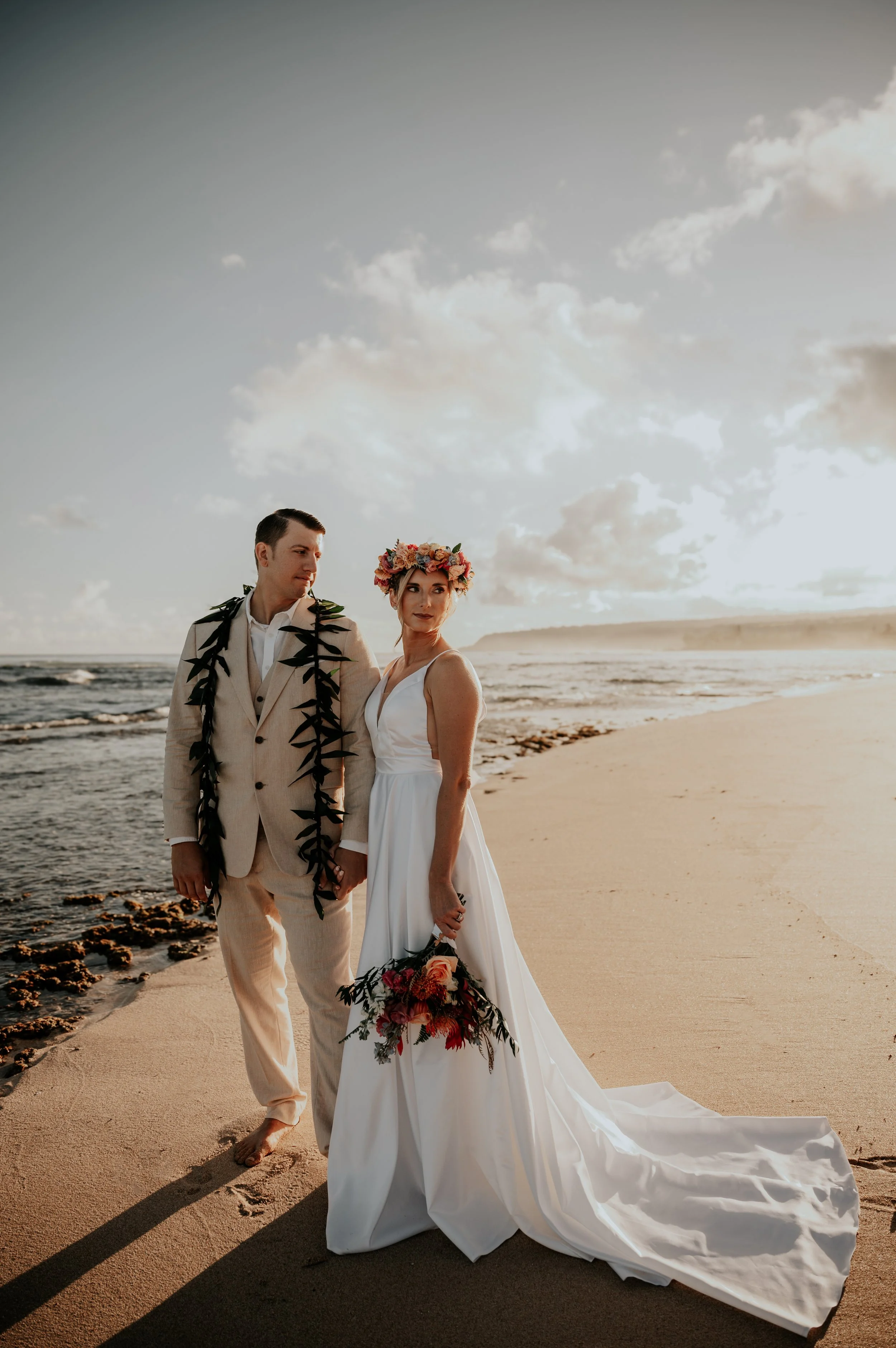 A bride and groom standing on a beach at sunset, holding hands and looking at each other. The bride wears a white gown and a flower crown, holding a bouquet, and the groom wears a beige suit with a dark green lei.