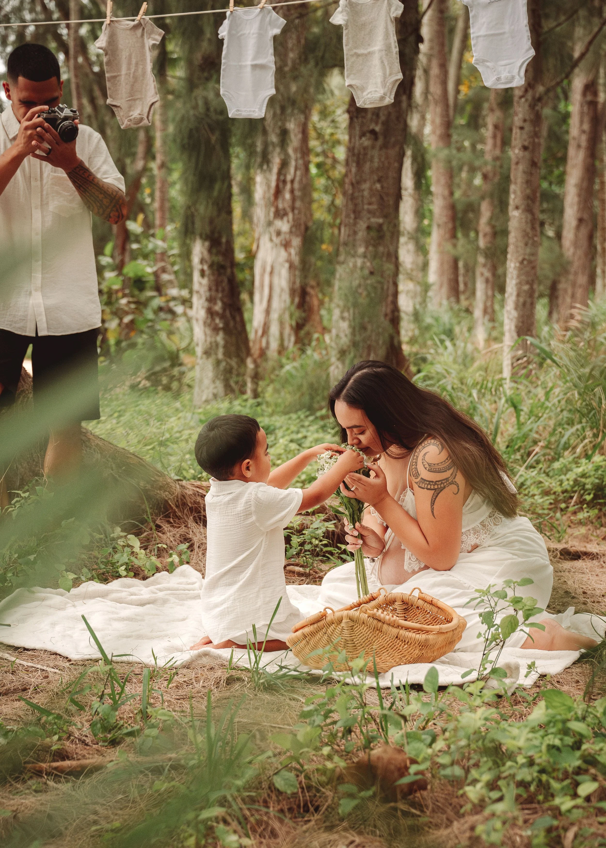A woman with tattoos and a boy sitting on a blanket in a forest, exchanging flowers. Clothes hang on a line above. A man nearby is taking a photo.