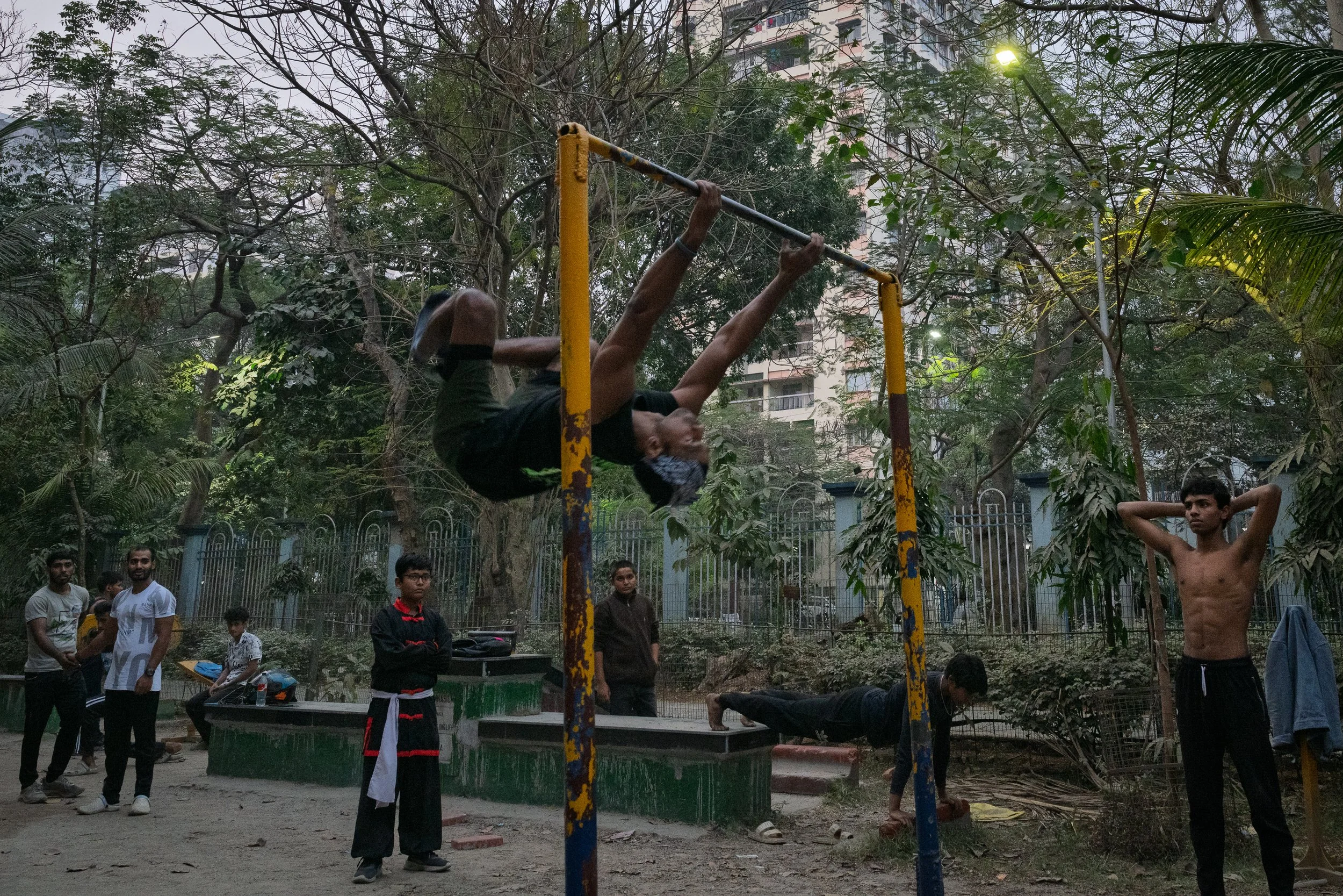 Young man in black shorts doing a pull-up on a rusty outdoor pull-up bar in a park, surrounded by trees and other young people, some watching and others exercising.