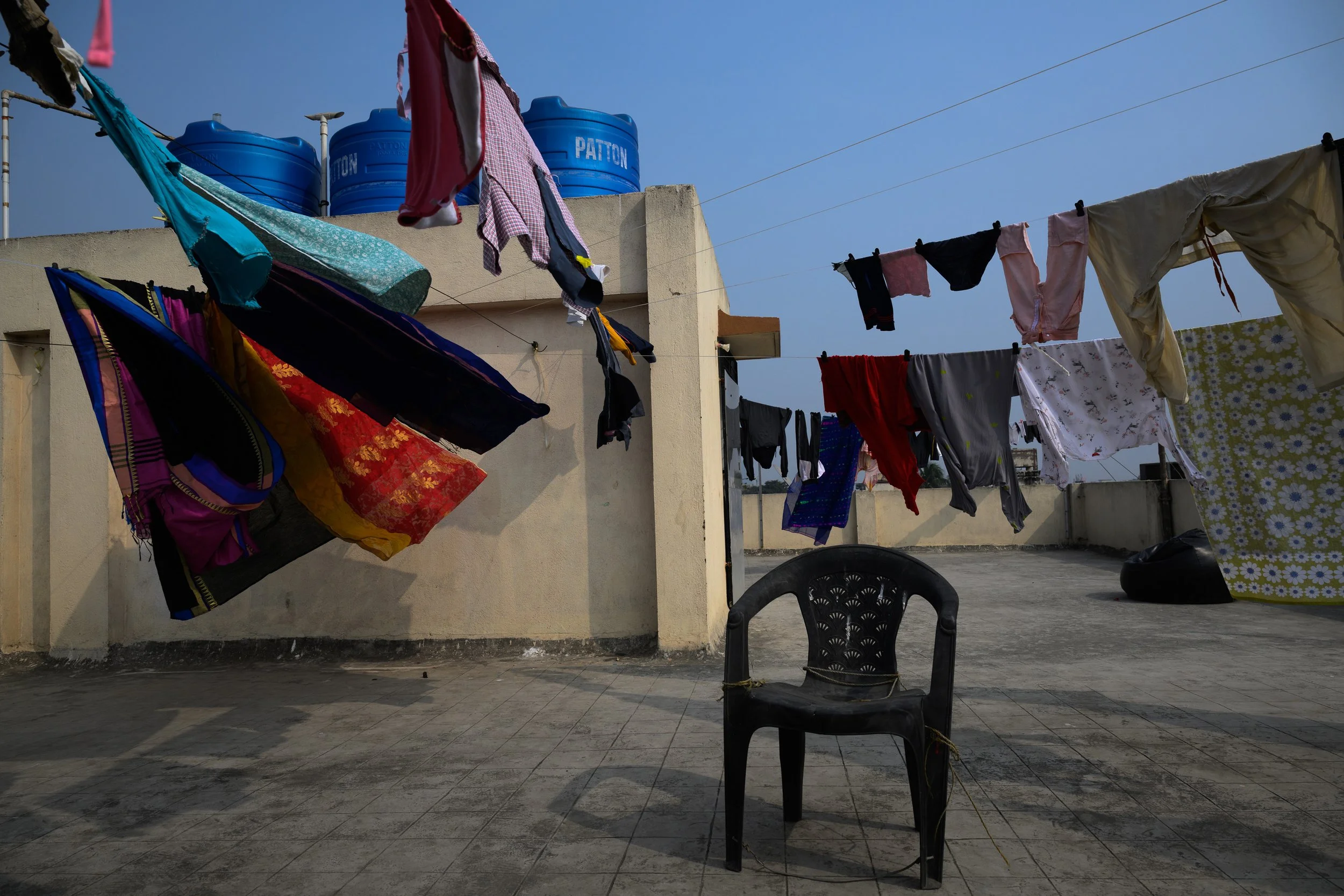 Clothes hanging on lines for drying on a rooftop with a black plastic chair in the foreground and water tanks on the roof in the background.