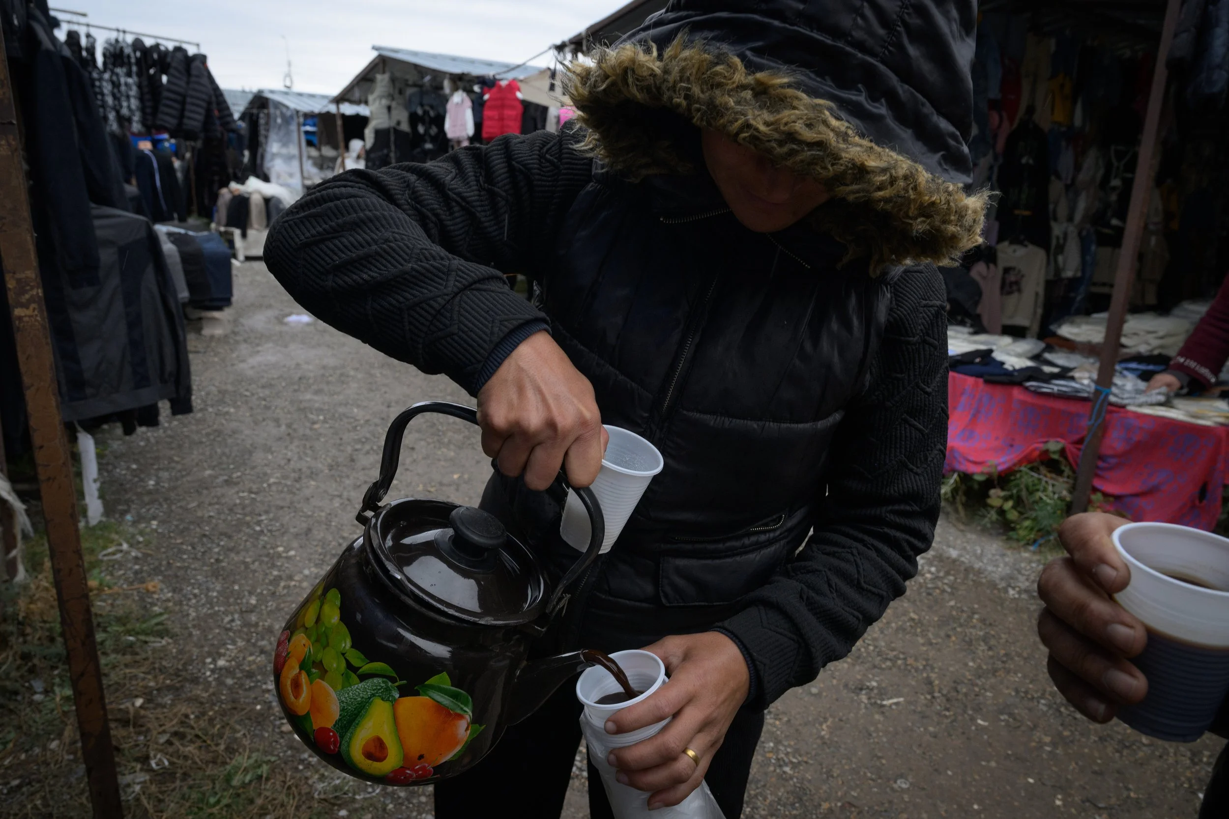 Person pouring a hot beverage from a colorful teapot into a paper cup at an outdoor market.