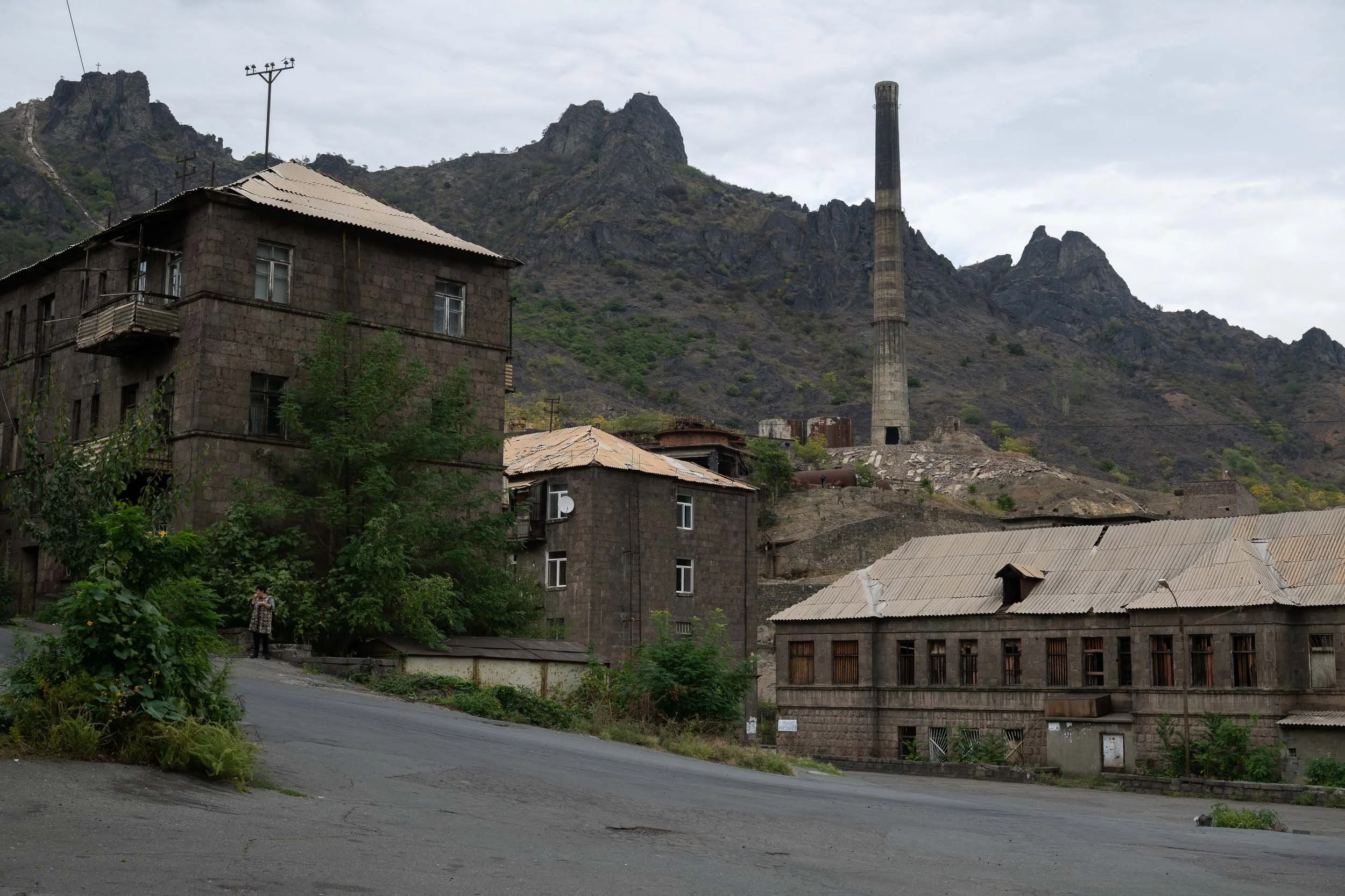 A view of an urban area with old stone buildings, lush greenery, and a mountain with rocky formations in the background under a cloudy sky.