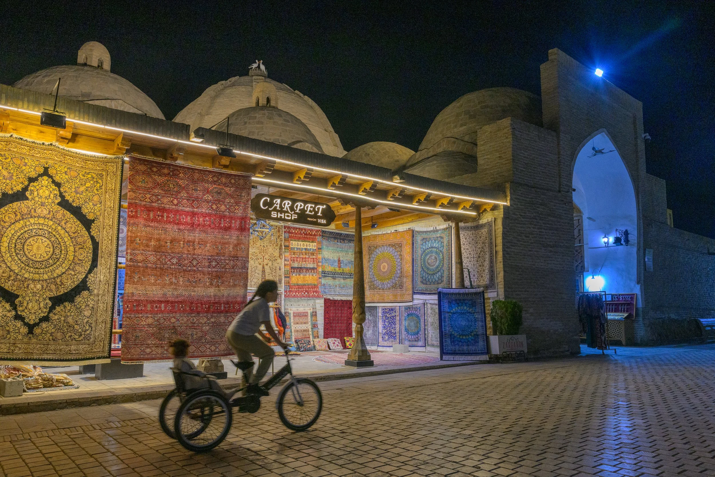 Night scene of outdoor carpet shop with hanging rugs, illuminated signage, a person riding a bicycle with a child, and traditional architectural elements in the background.