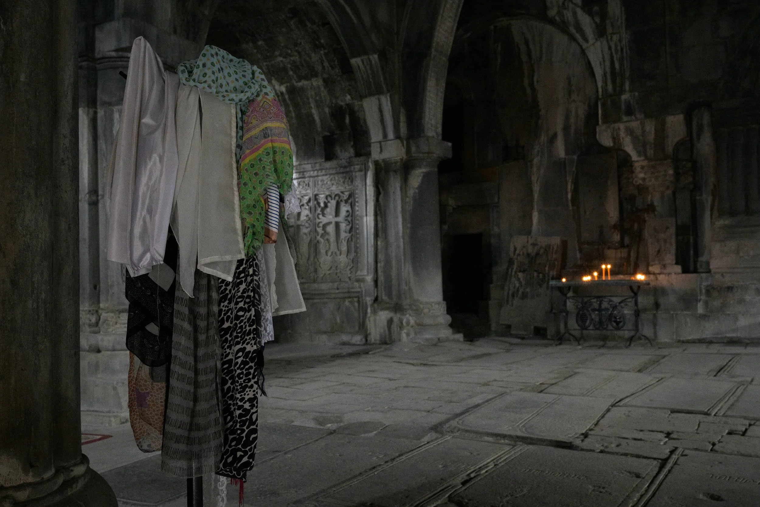 A display of various colorful fabrics hanging inside an ancient stone structure with ornate carvings and lit candles in the background.