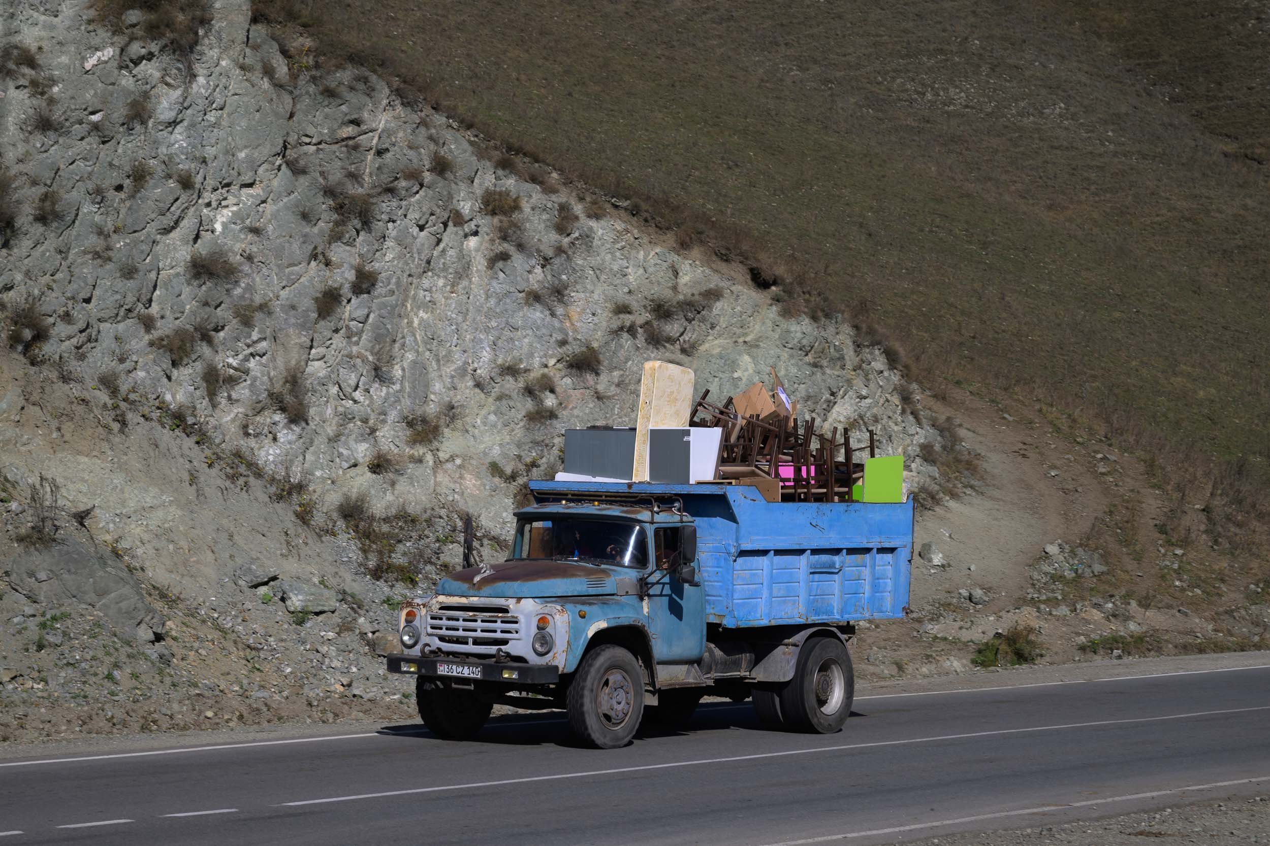 An old, blue flatbed truck driving on a highway beside a rocky hillside with sparse vegetation.
