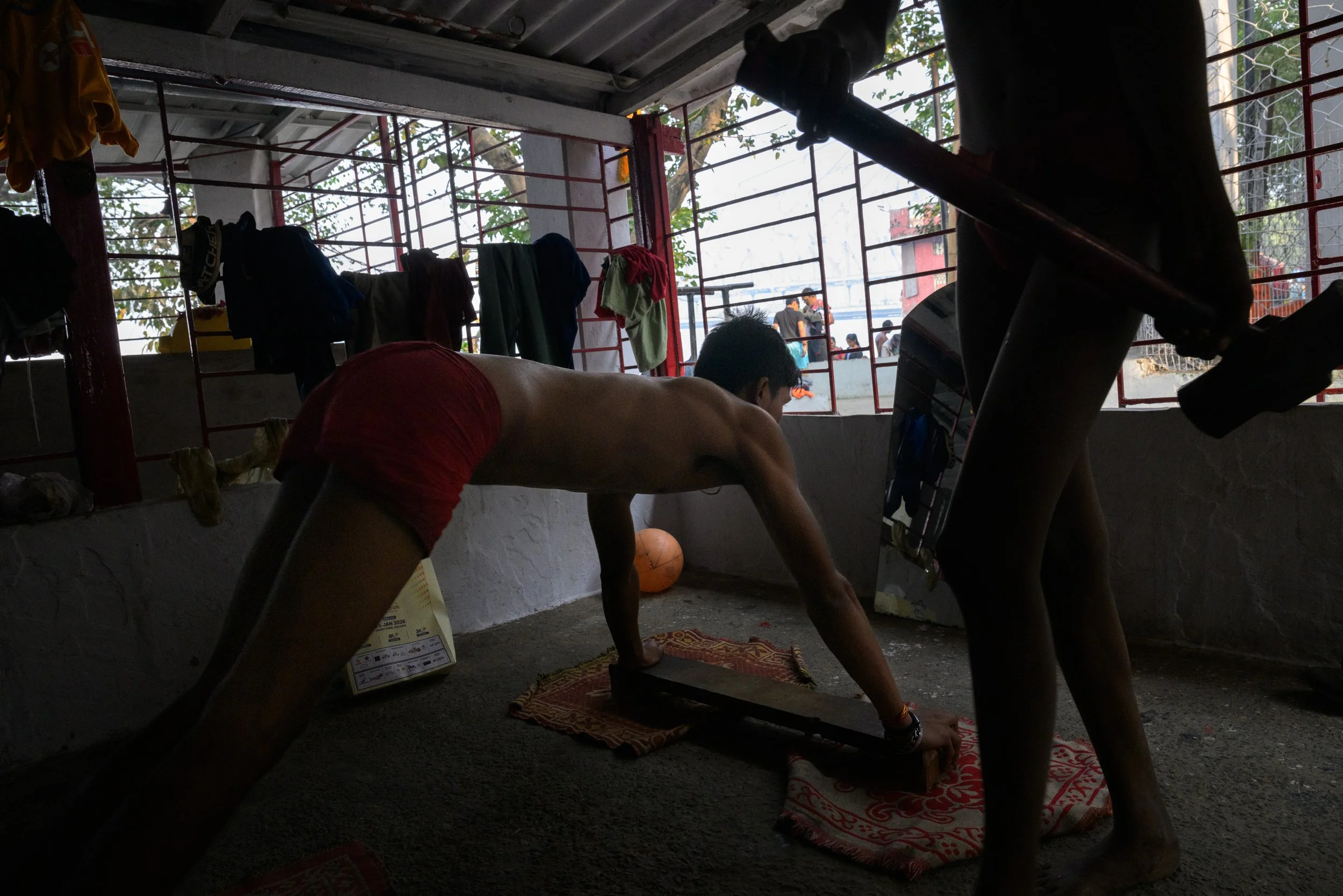 A young man performing a push-up on a wooden plank in a rustic indoor gym with laundry hanging from the ceiling and spectators outside the window.