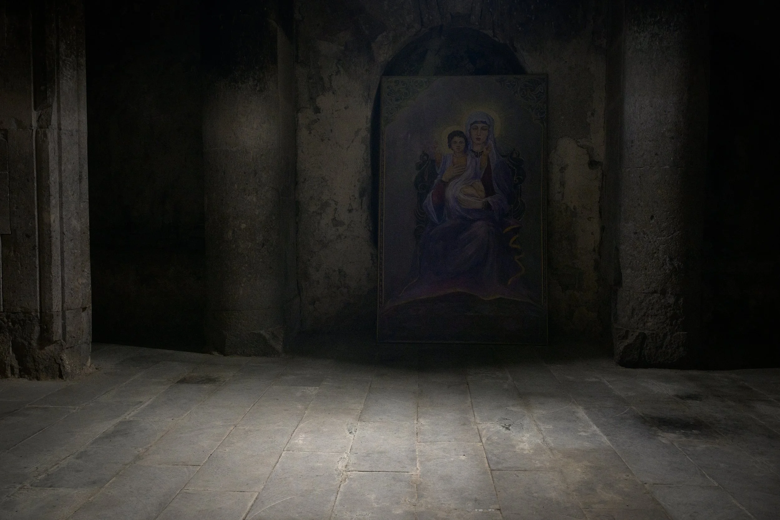 A religious painting of the Virgin Mary holding baby Jesus, set against a dark, stone interior with stone columns and a stone floor.