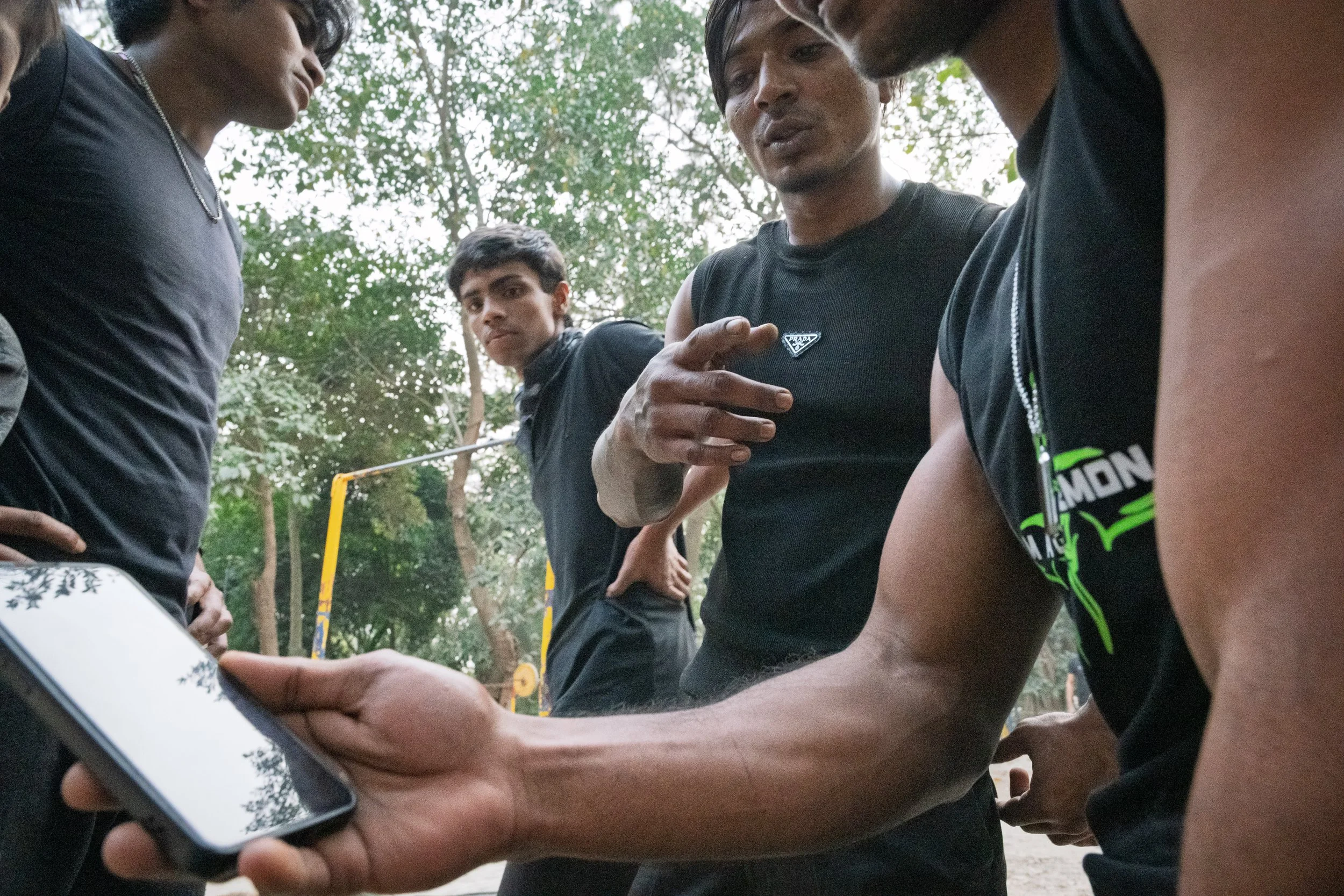 A group of young men outdoors, one showing something on a smartphone to others, with trees in the background.