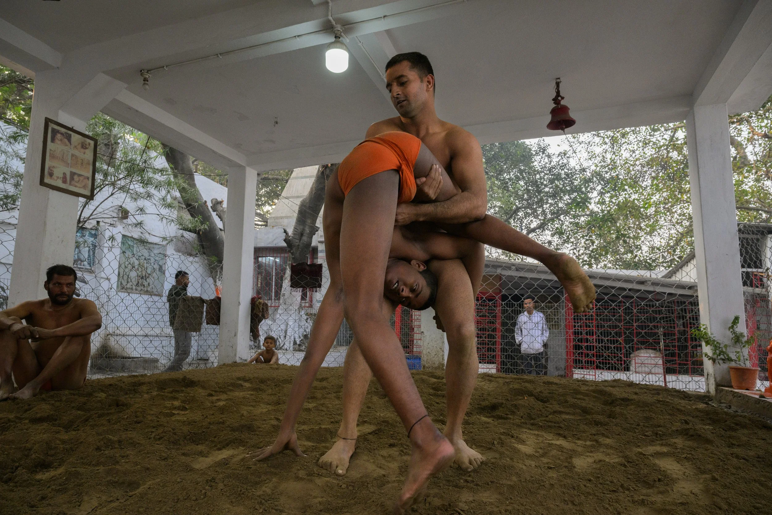 Two men are engaged in a traditional wrestling match inside a covered courtyard, with onlookers observing nearby.