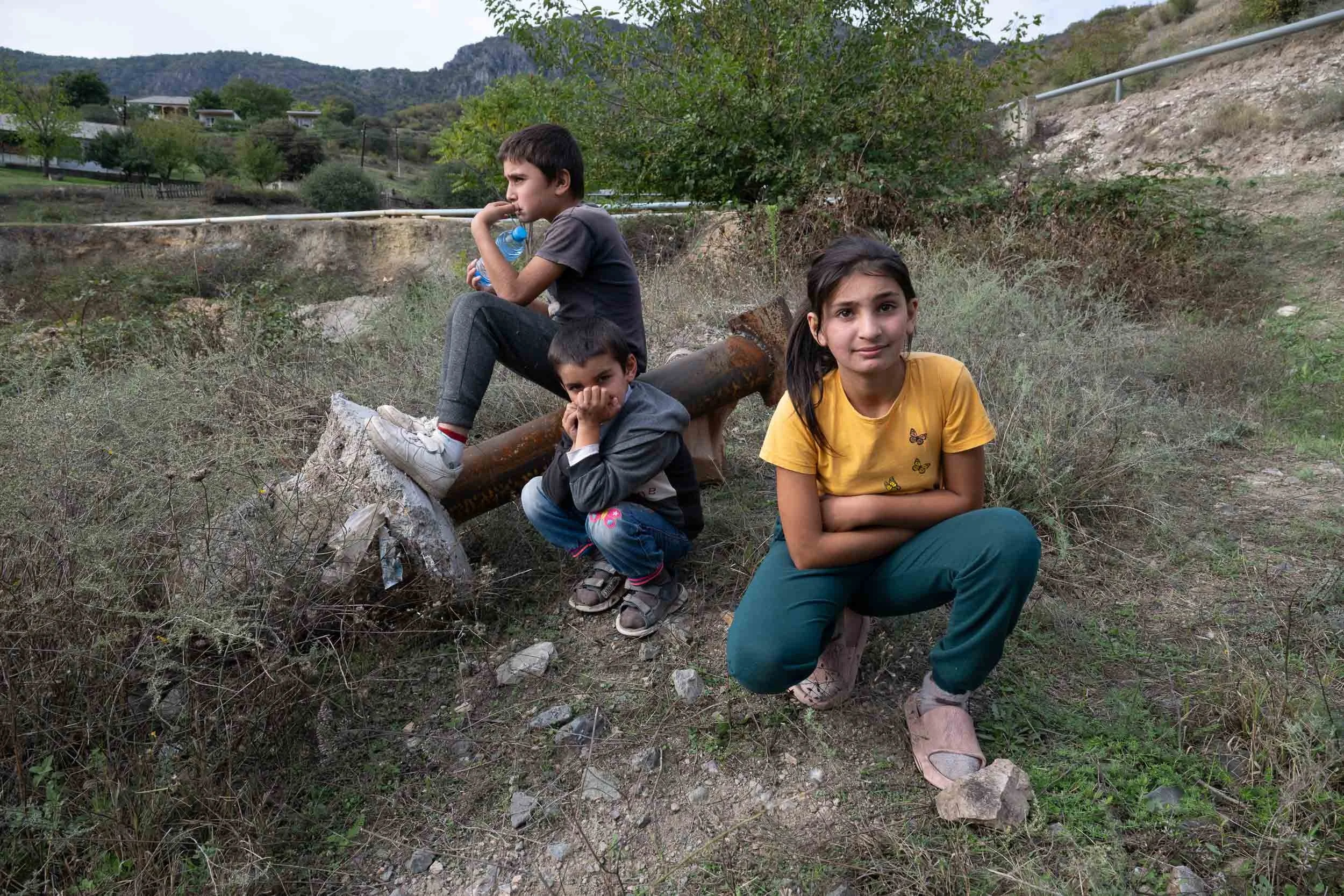Three children sitting outdoors on grass and dirt in a rural area with hills, trees, and mountains in the background. One boy is holding a water bottle and looking to the side, while a girl in a yellow shirt and another boy with a finger in his mouth