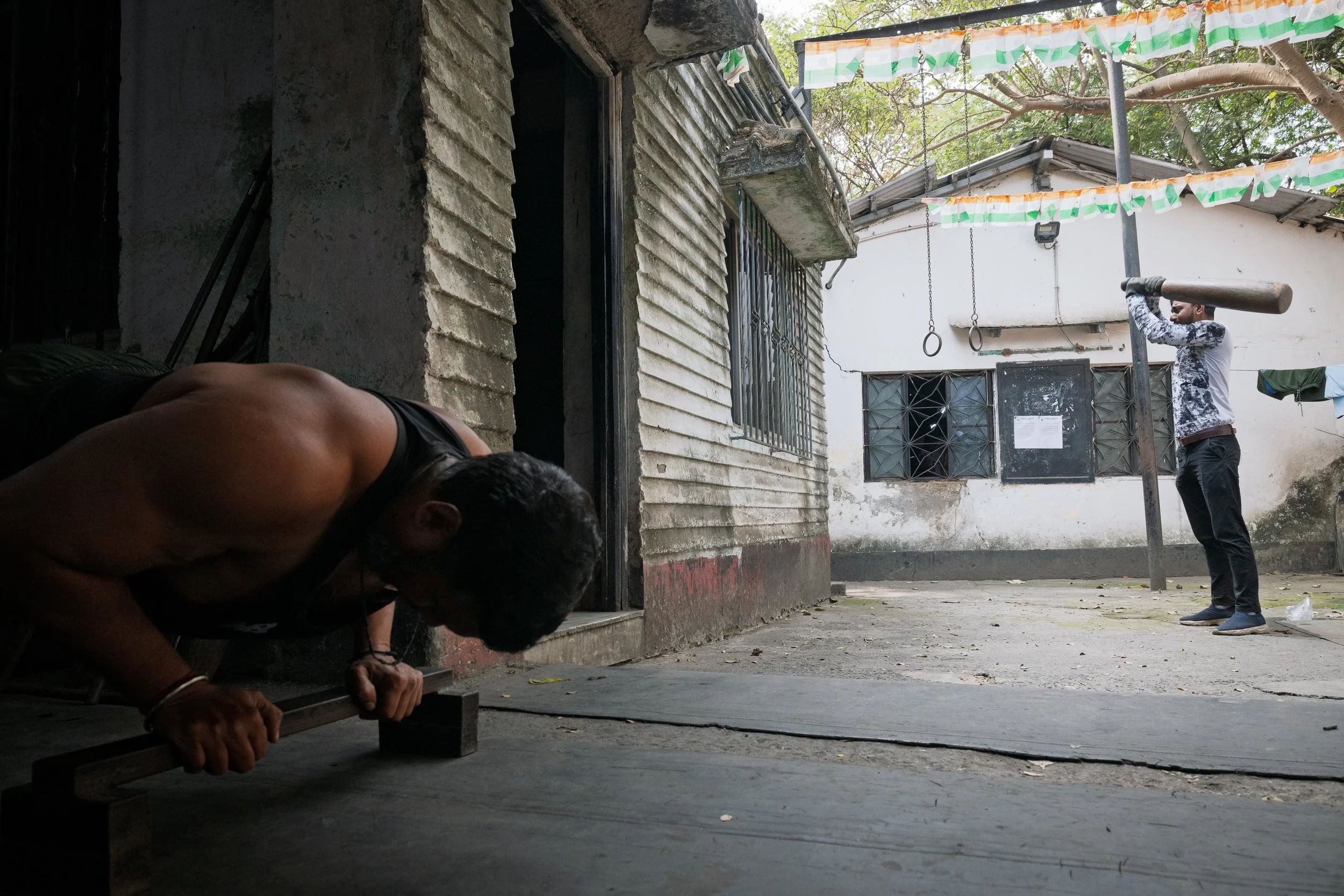 A man doing push-ups near a brick wall and an open doorway, while another man using a wooden log as a barbell stands outdoors in a yard, with trees and a building in the background.