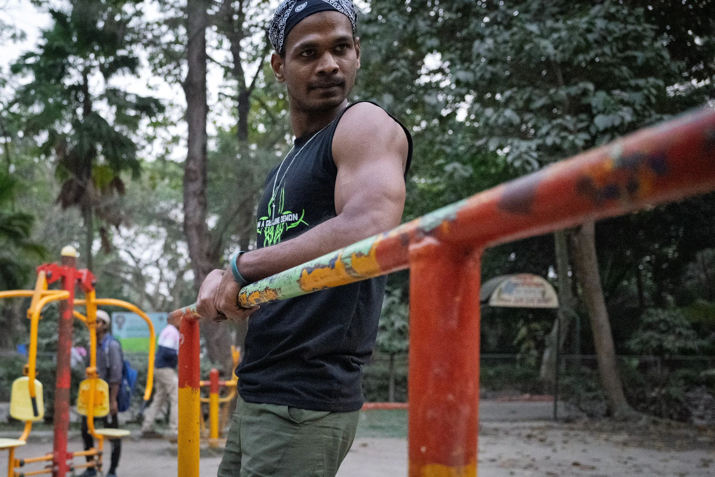 A young man in a black sleeveless shirt and bandana standing at an outdoor gym, holding a rusted orange and green bar, with other exercise equipment and people in the background.