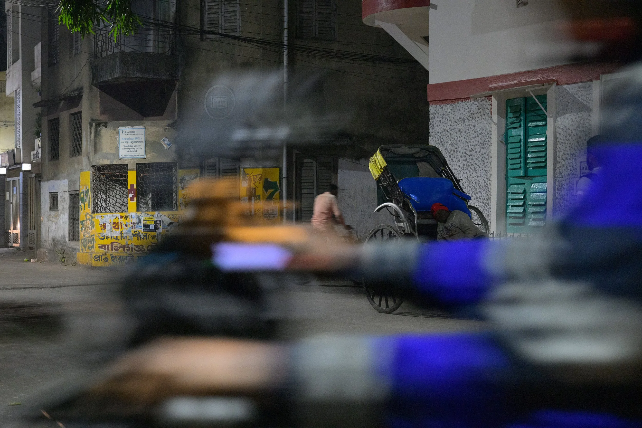 Street scene at night with a rickshaw, a person writhing on the ground, a woman walking, lit by streetlights and smoke in the background.