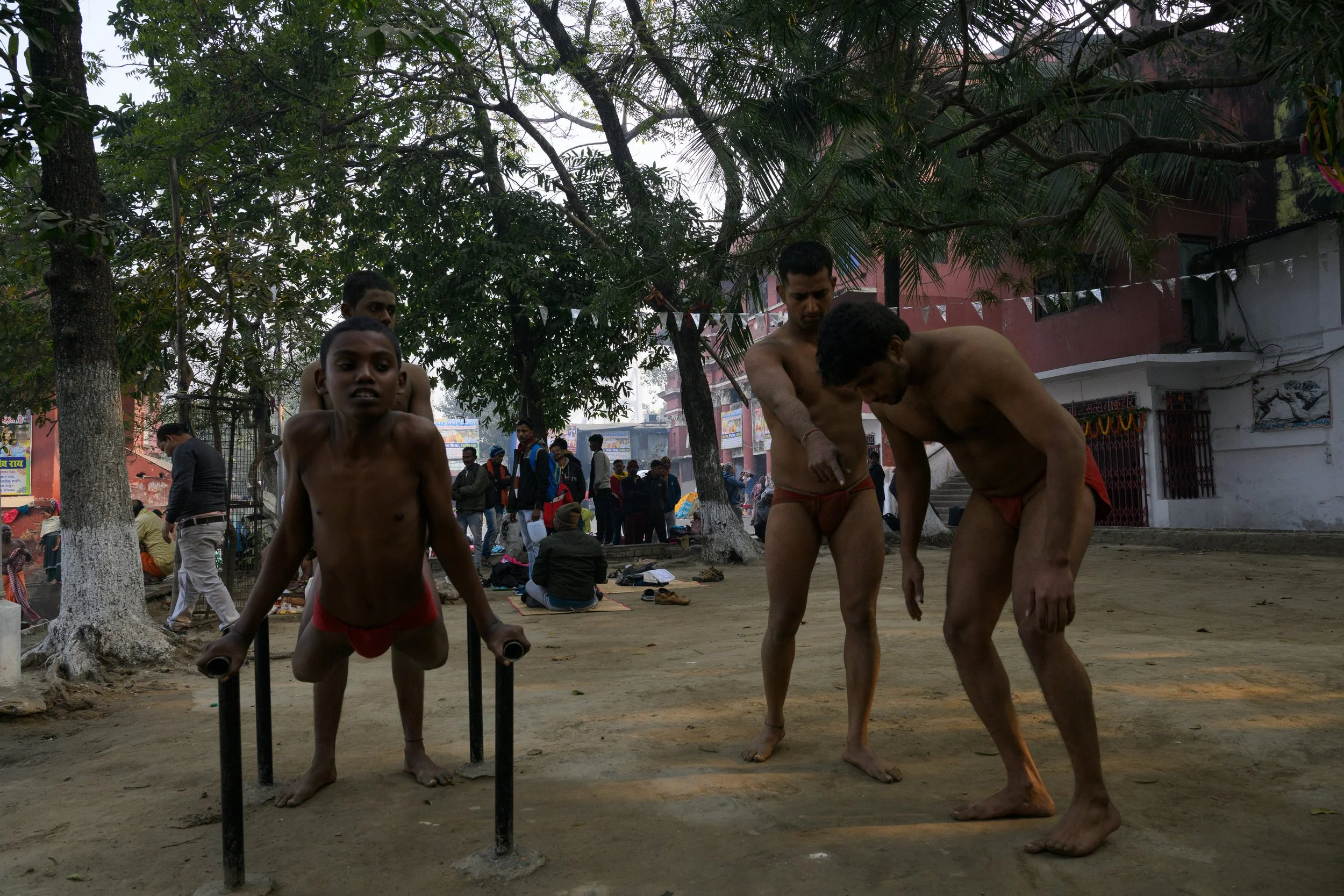Men and boys in traditional loincloths practicing wrestling outdoors under trees, with spectators watching in the background.