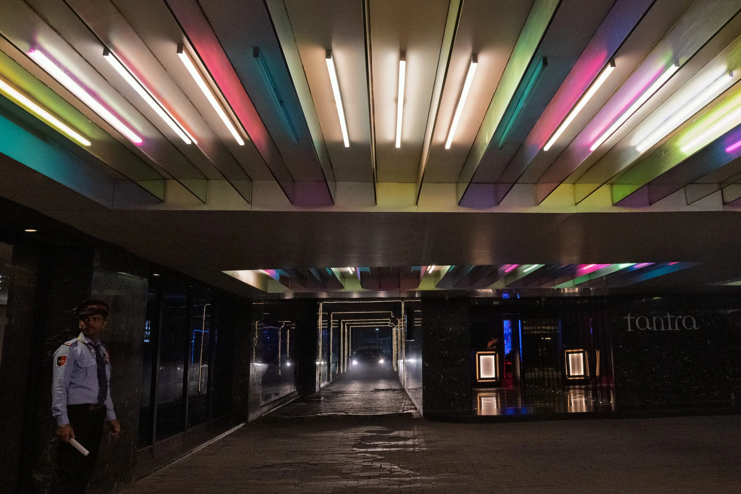Nighttime scene of a modern building entrance with colorful neon lights on the ceiling, a security guard standing to the left, and a dark, reflective corridor leading into a well-lit interior space.