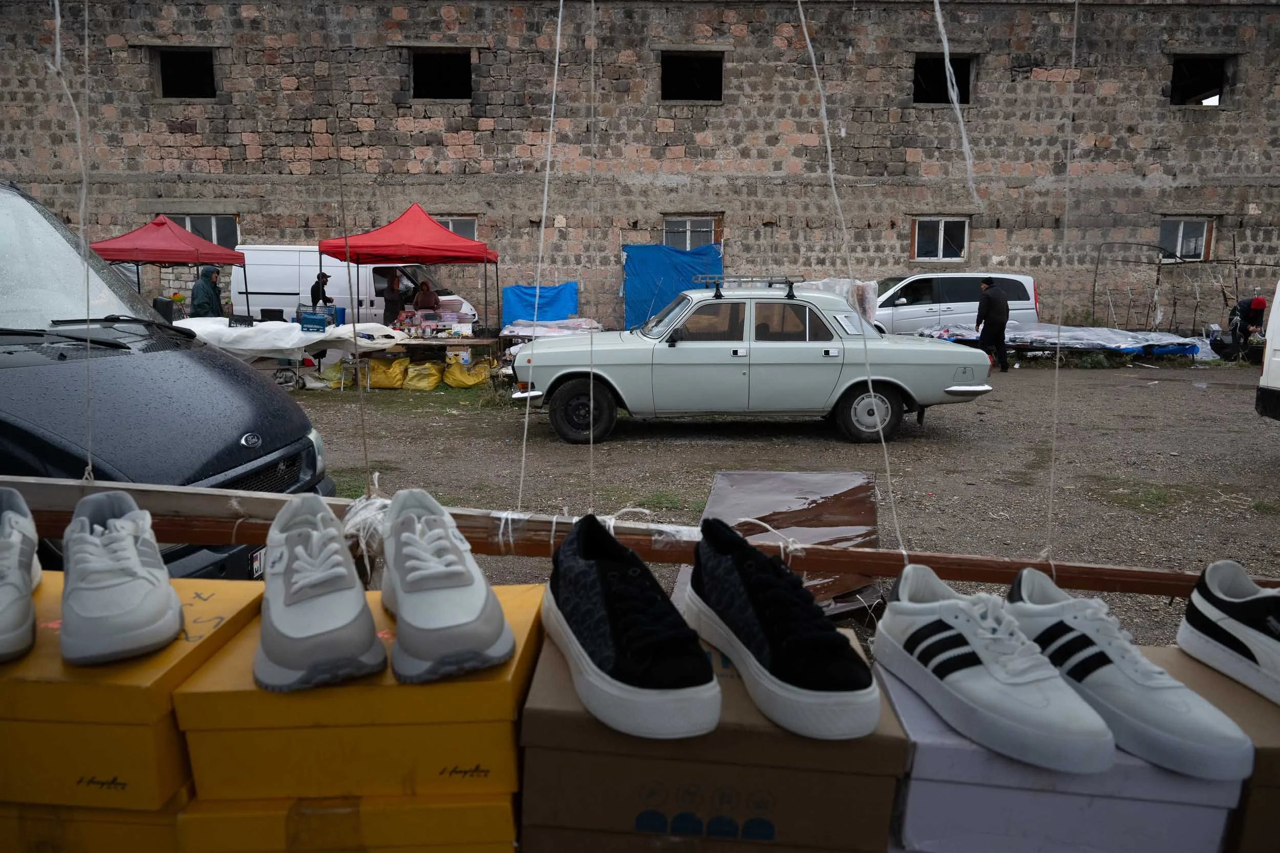 A shoe stand with white sneakers displayed in front of an outdoor market scene, featuring vendors with red tents, parked vehicles, and a brick wall in the background.