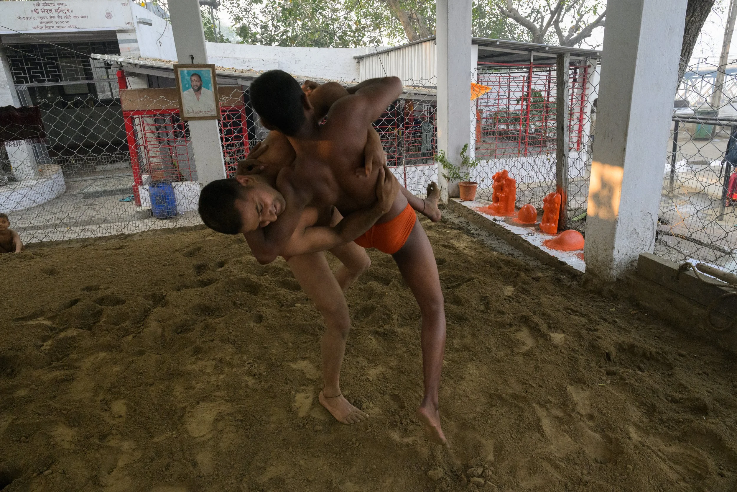 Two young men practicing wrestling on a dirt floor in a confined space, with one lifting the other. There are decorative orange objects and potted plants on the side, and a wire fence in the background.