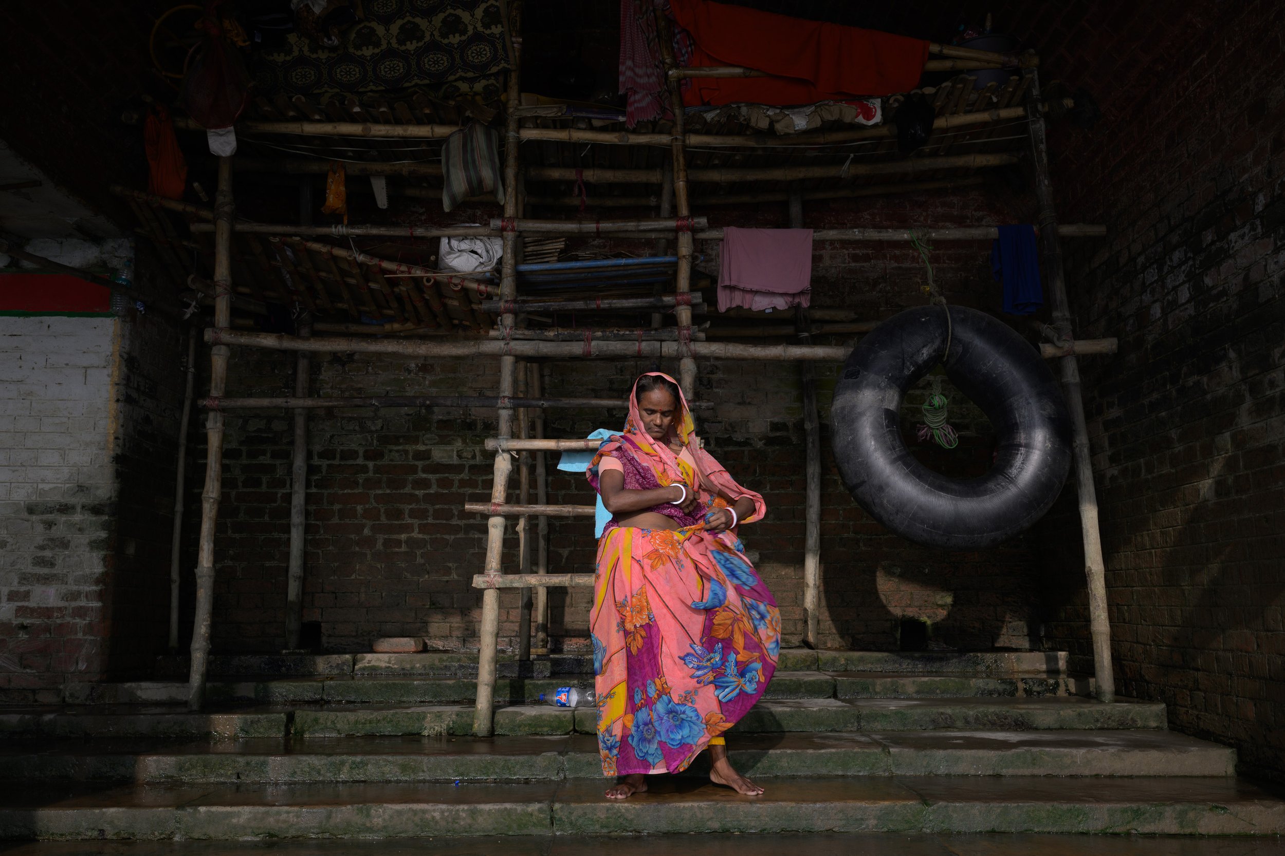 A woman in a colorful saree sitting on concrete stairs under a bamboo scaffold structure in a dimly lit area with brick walls, a black rubber tire hanging, and laundry hanging overhead.