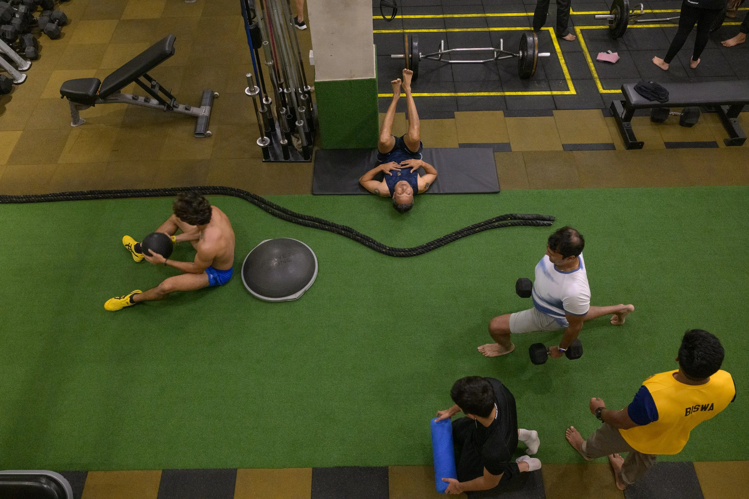 People exercising at a gym with various equipment, some performing different workouts on a green mat area.