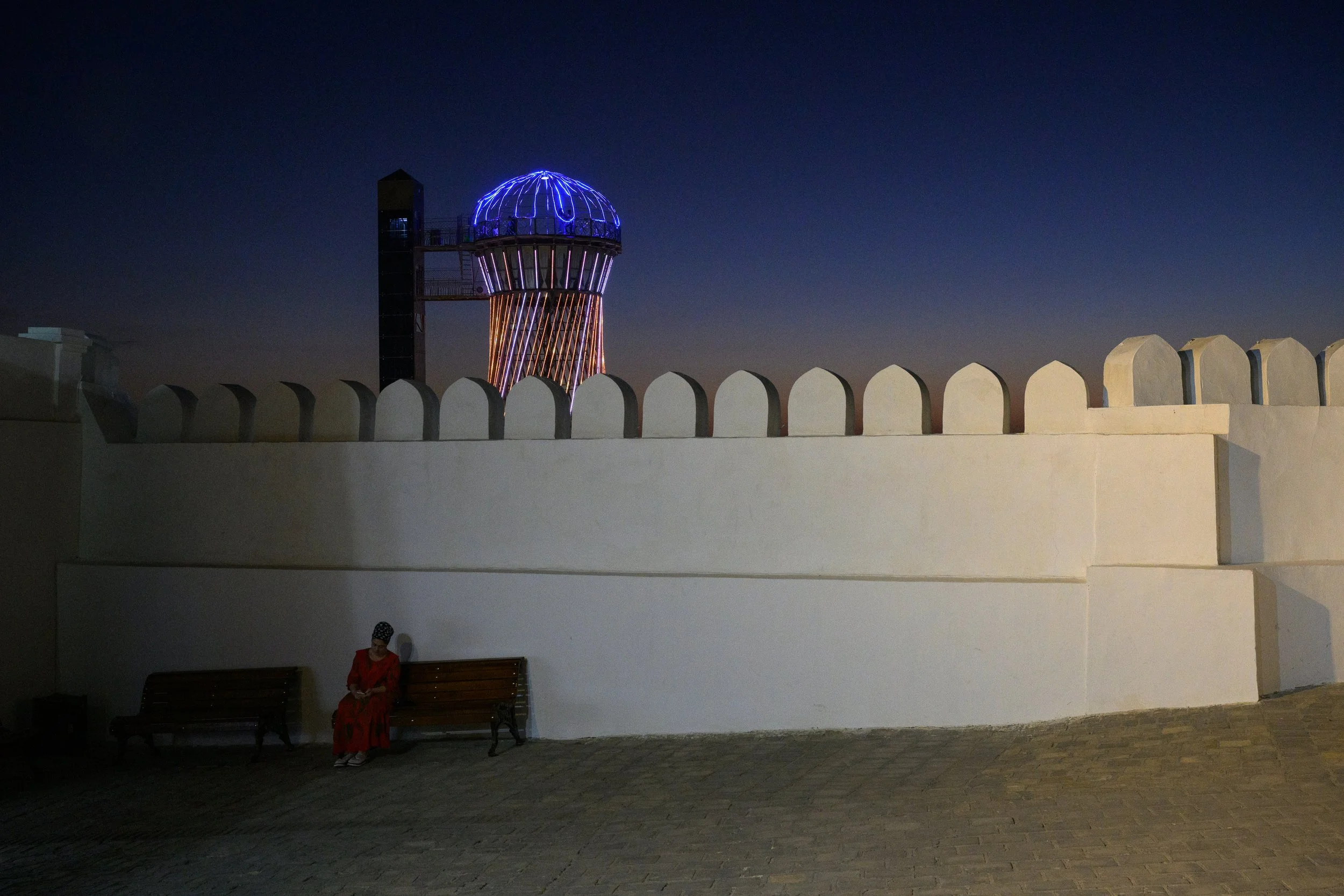 A woman in a red dress and headscarf sitting on a bench in front of a white wall with a decorative top, with a modern tower with colorful lights illuminated against a dark night sky in the background.
