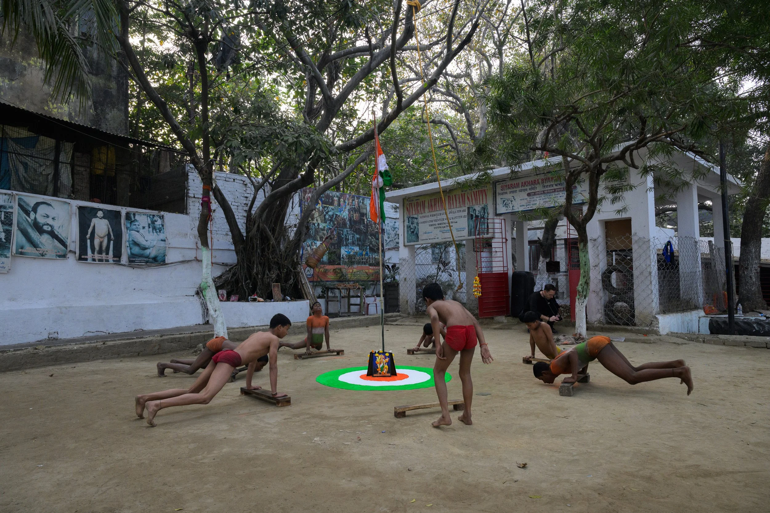 Young boys practicing traditional Indian game of flying kites in an outdoor courtyard with trees, banners, and posters on the walls.
