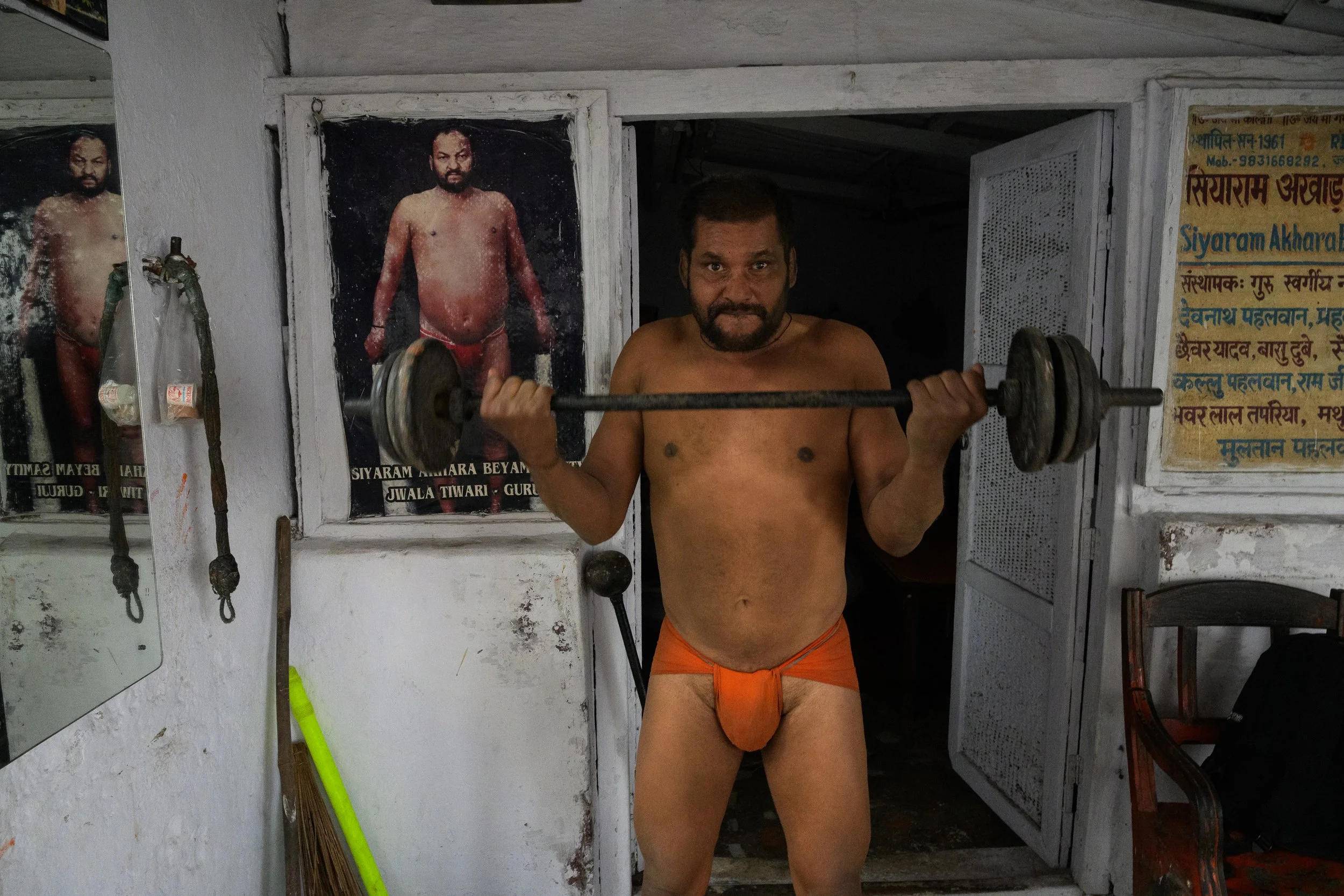 A man lifting a barbell inside a room with posters of a spiritual figure on the wall. The man is shirtless, wearing orange clothing, and standing in front of an open door.