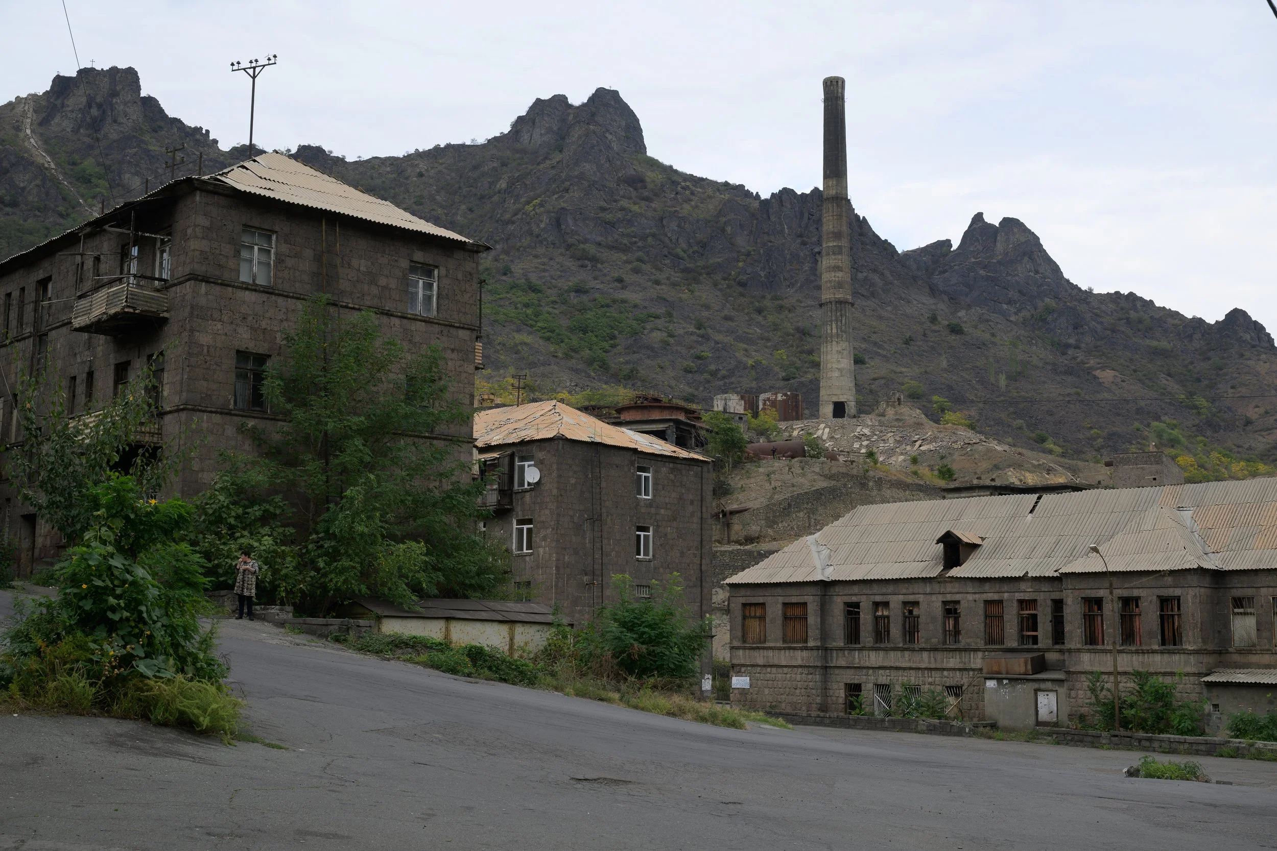 A small town at the foot of a mountain range with a tall industrial chimney, old stone buildings, trees, and overcast sky.