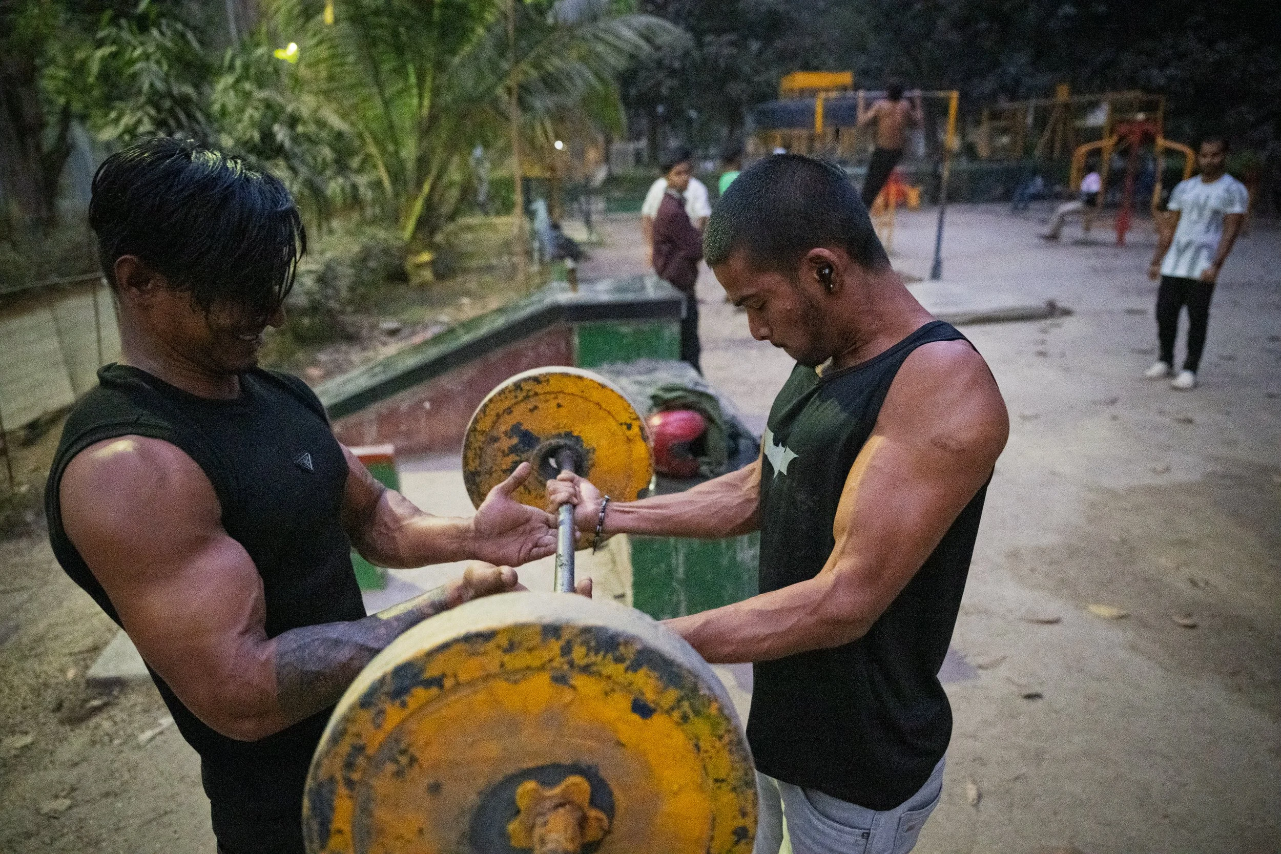 Two muscular young men with short dark hair workout with a barbell at an outdoor gym during evening. One man, wearing a sleeveless black shirt, is spotting the other, who is also in a sleeveless black shirt, lifting the barbell. In the background, ot