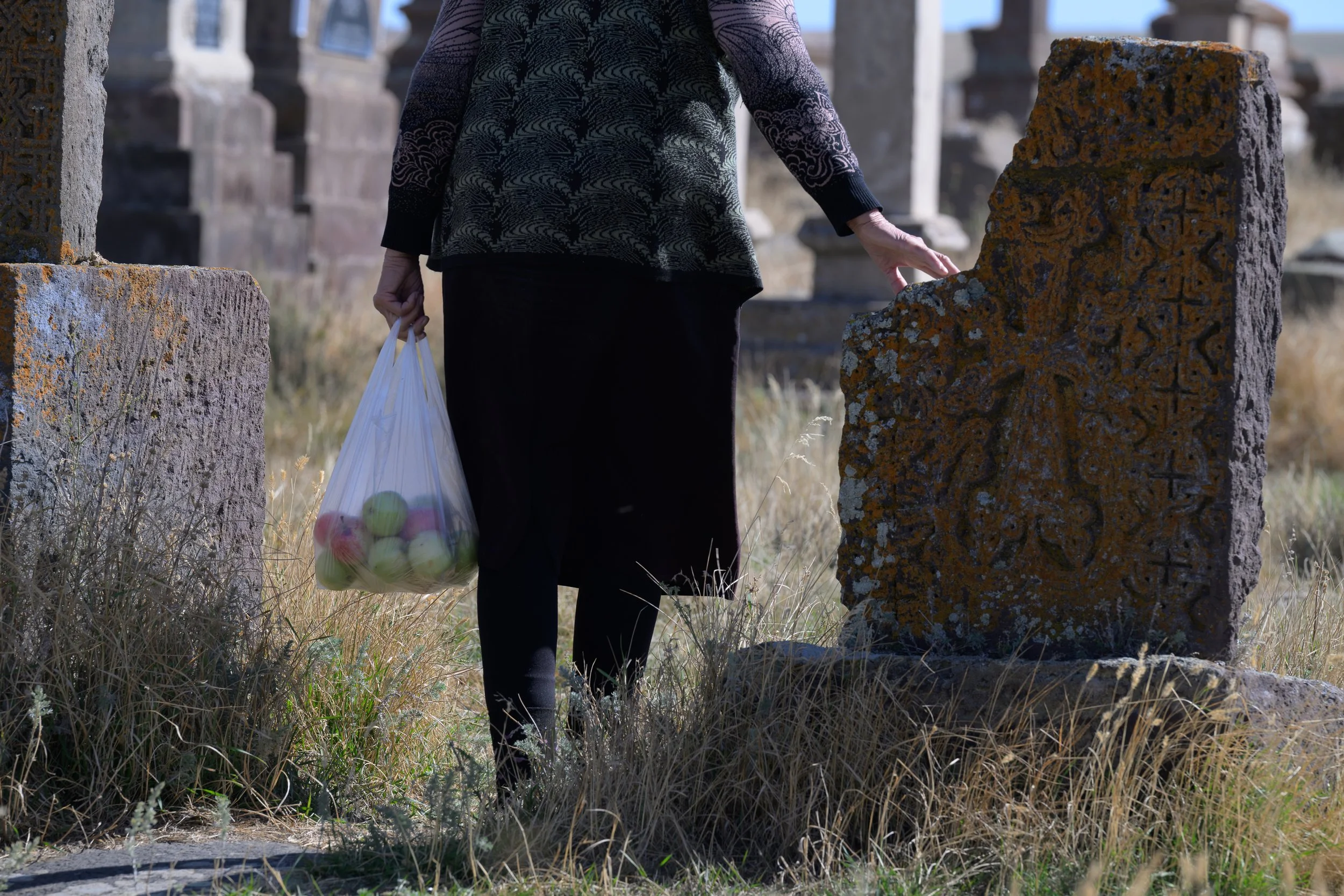 Person wearing a black skirt and a patterned sweater touching an old, moss-covered gravestone in a cemetery, holding a bag of apples.