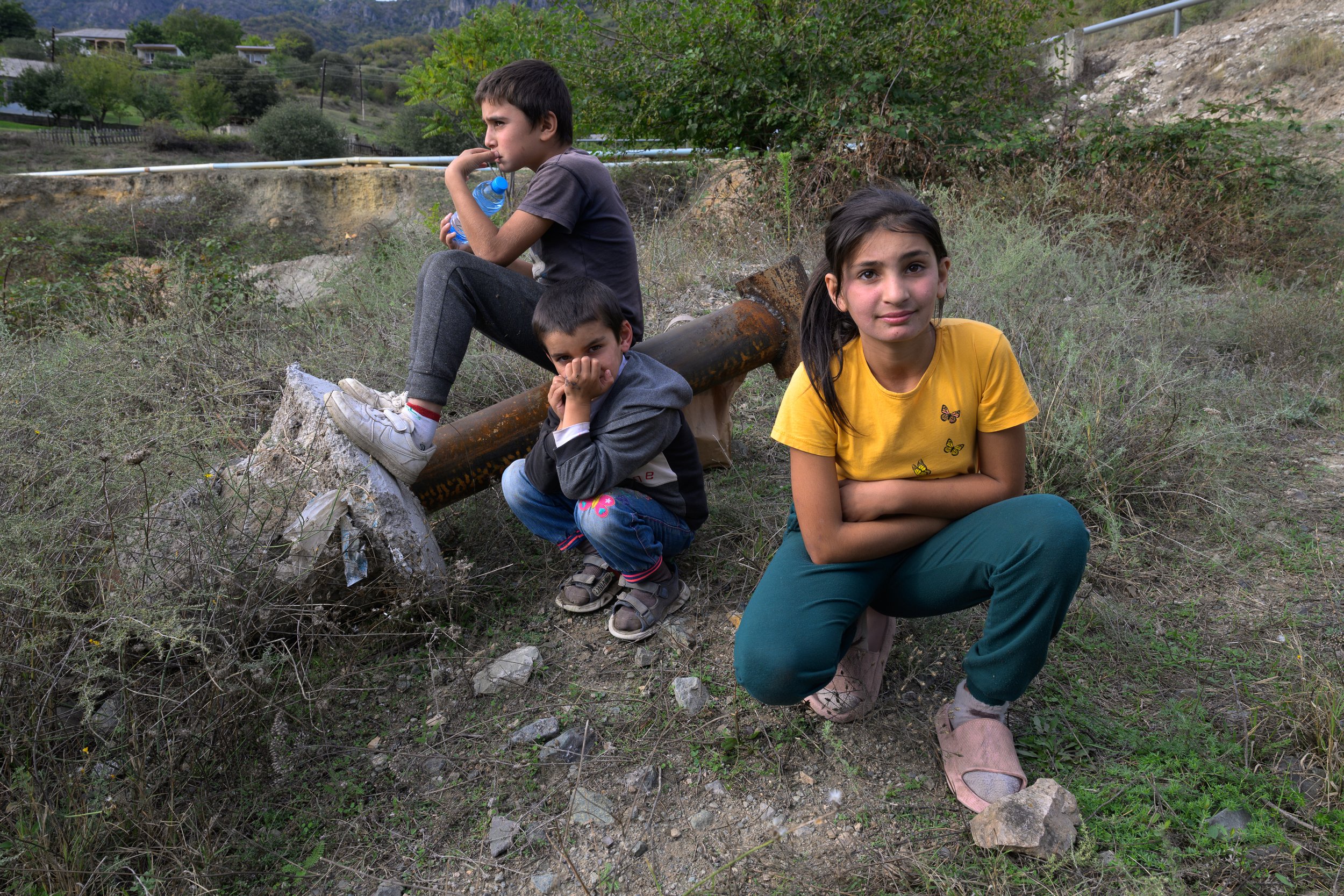 Four children sitting outdoors next to a rusty pipe, surrounded by dry grass and shrubs, with a hillside and trees in the background.