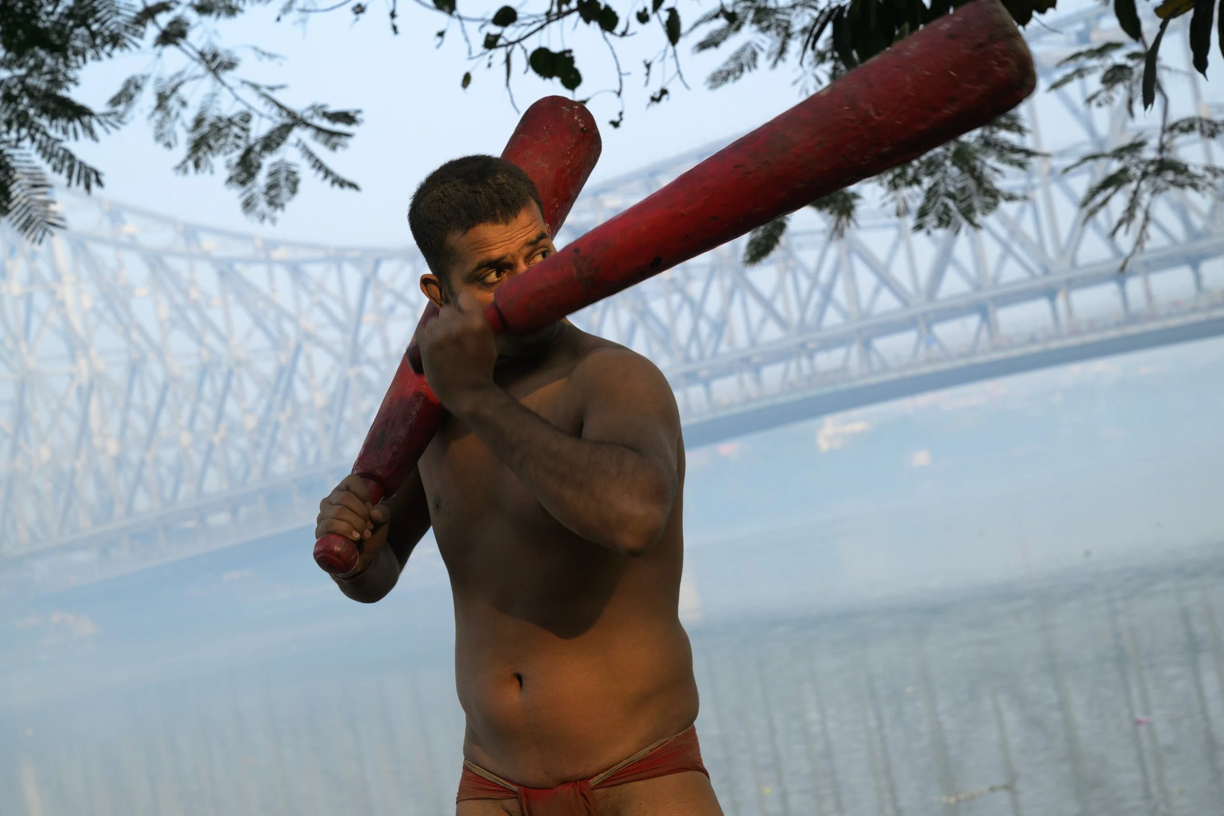 A man holding a large red wooden stick on his right shoulder, shirtless, standing near water with a bridge in the background and tree branches overhead.